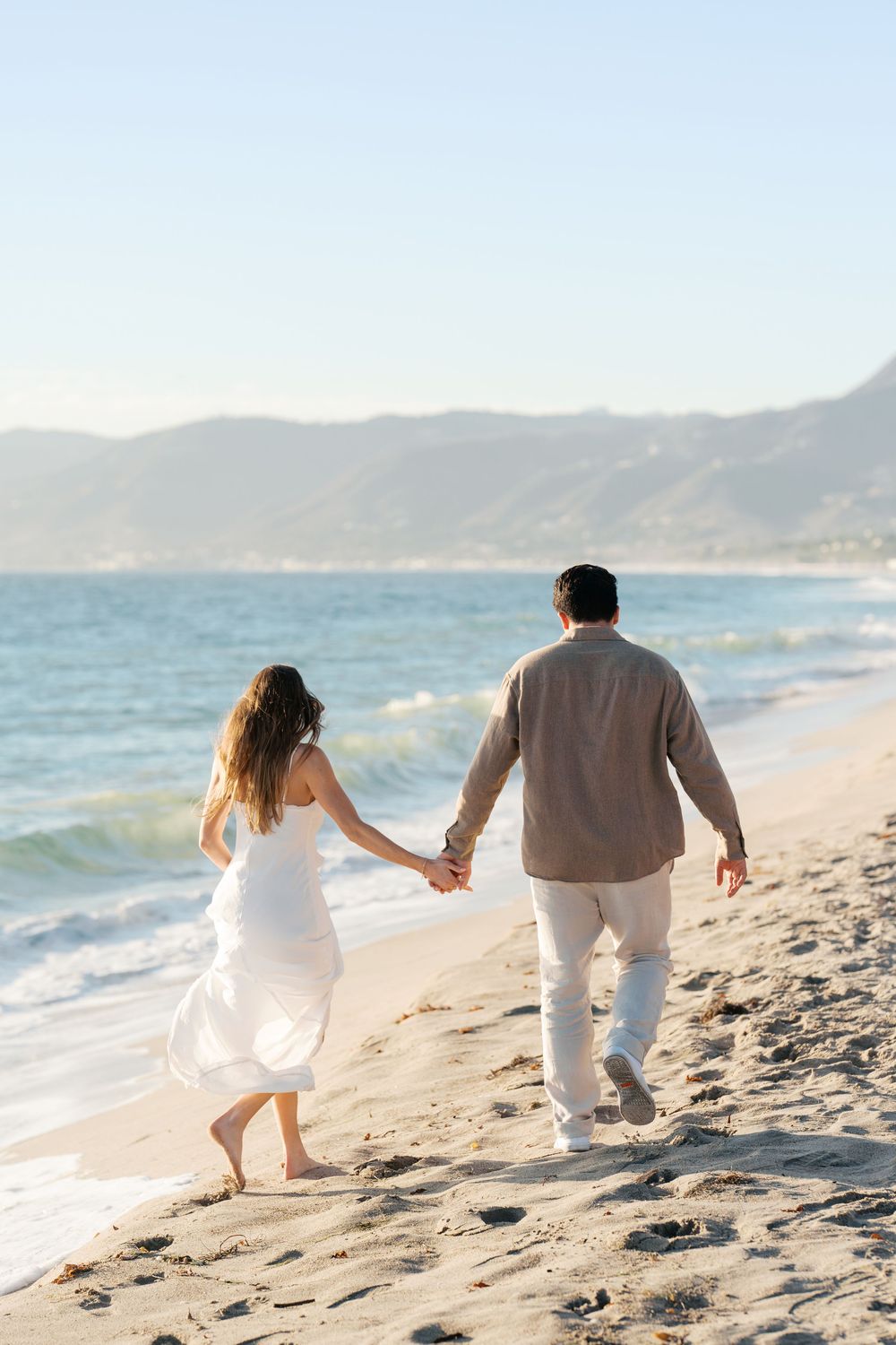 Couple walking hand in hand along a scenic beach with mountains and waves in the background at sunset.