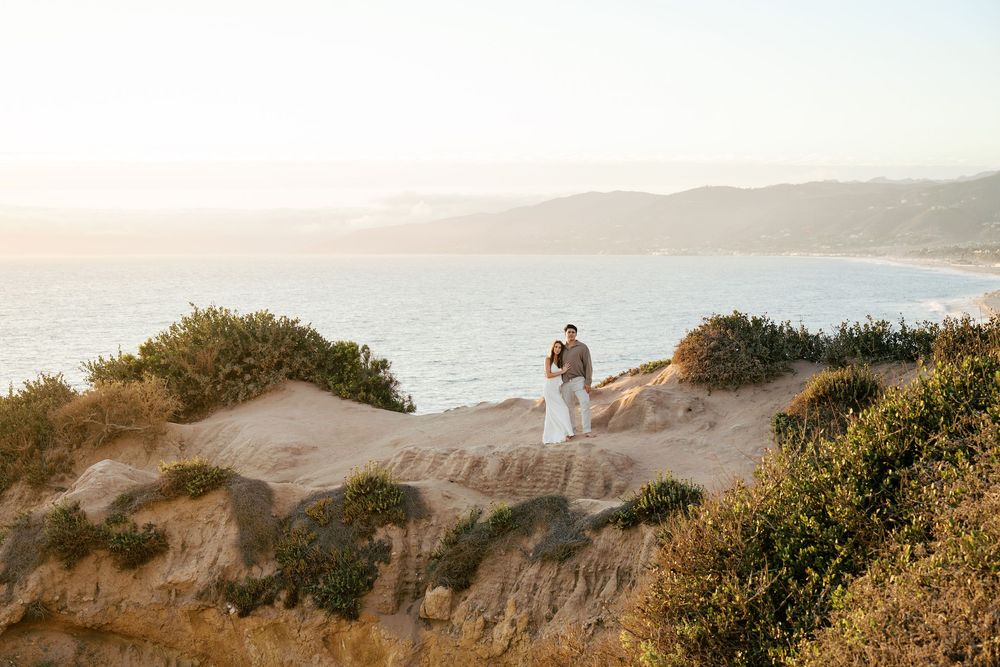 Couple walks along scenic coastal trail with golden hills and ocean views during sunset in California.
