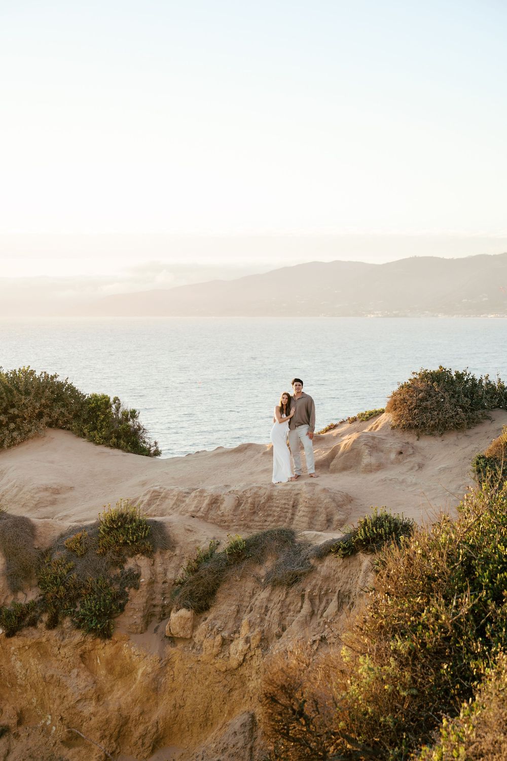 A couple stands on sandy cliffs overlooking the ocean at sunset with coastal vegetation in the foreground.