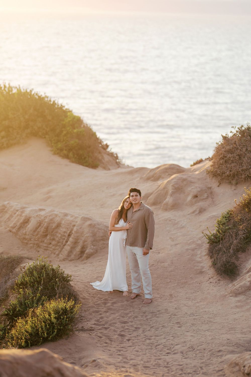 A couple embracing on coastal sand dunes at golden hour with the ocean in the background.