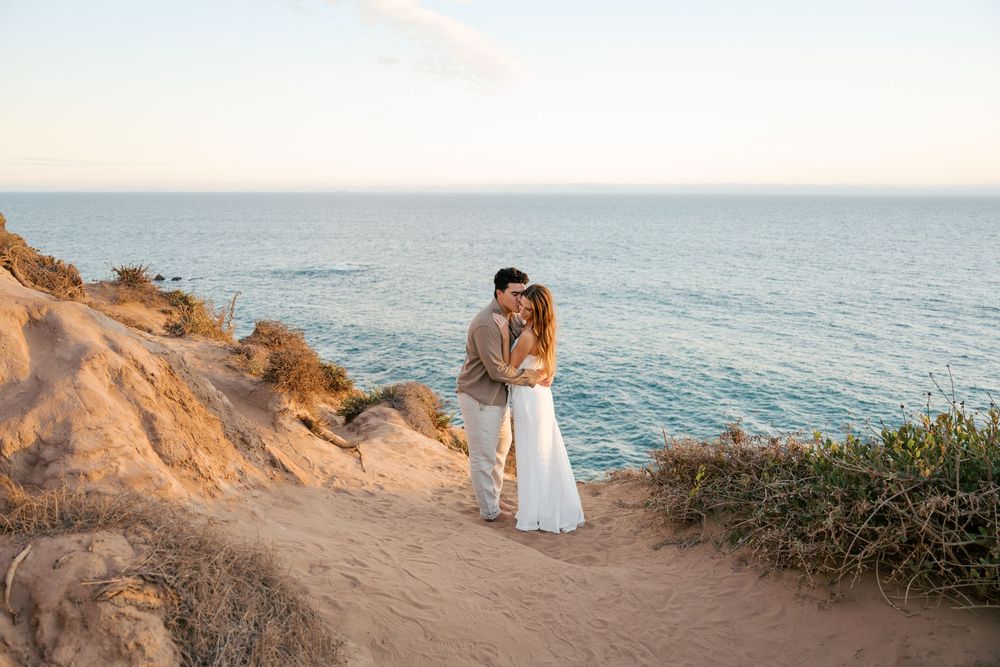 A couple in white outfits embraces on a scenic coastal cliff at sunset overlooking the ocean.