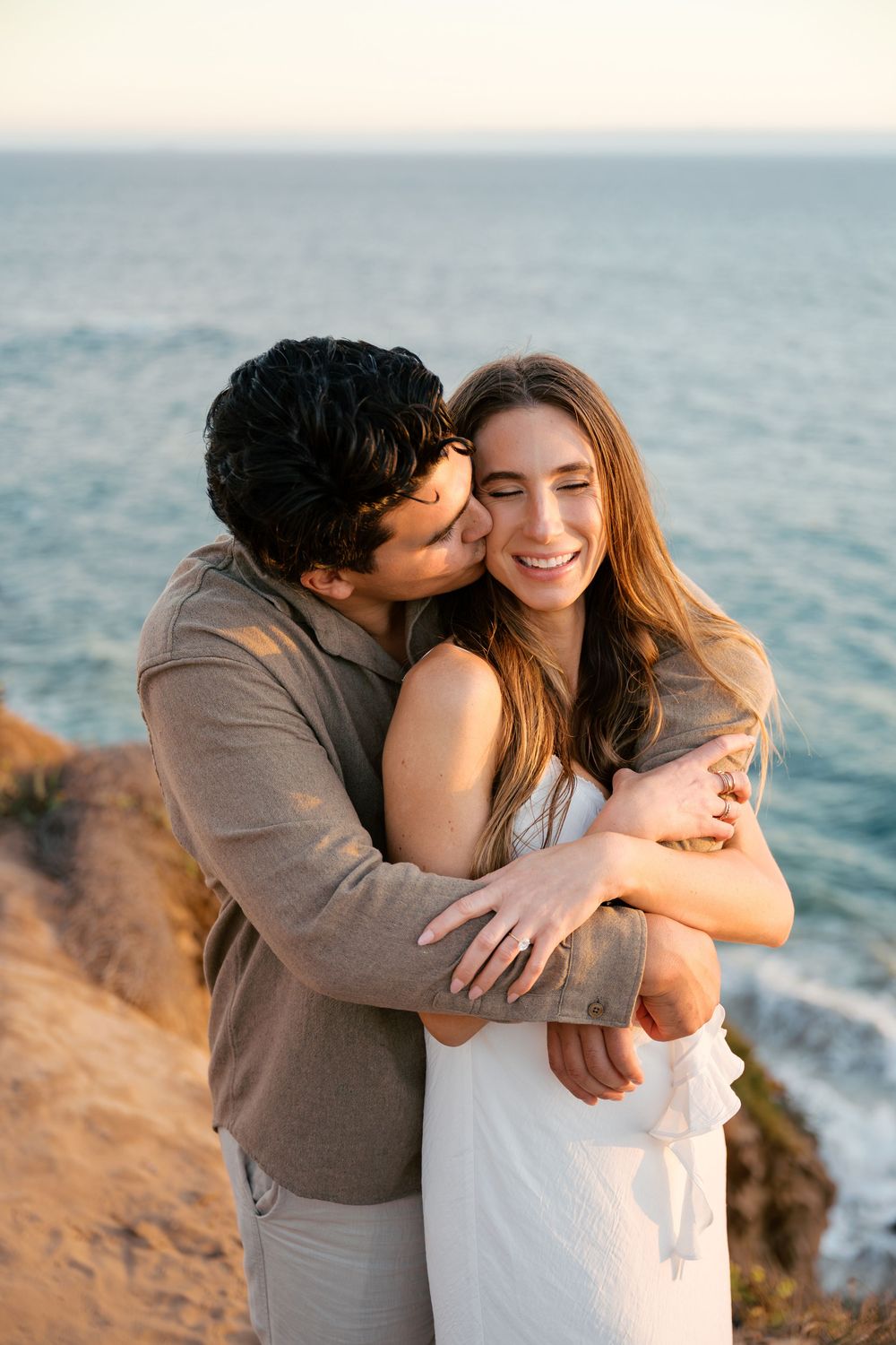 Romantic couple embraces on coastal cliffs during golden hour with ocean views in the background.