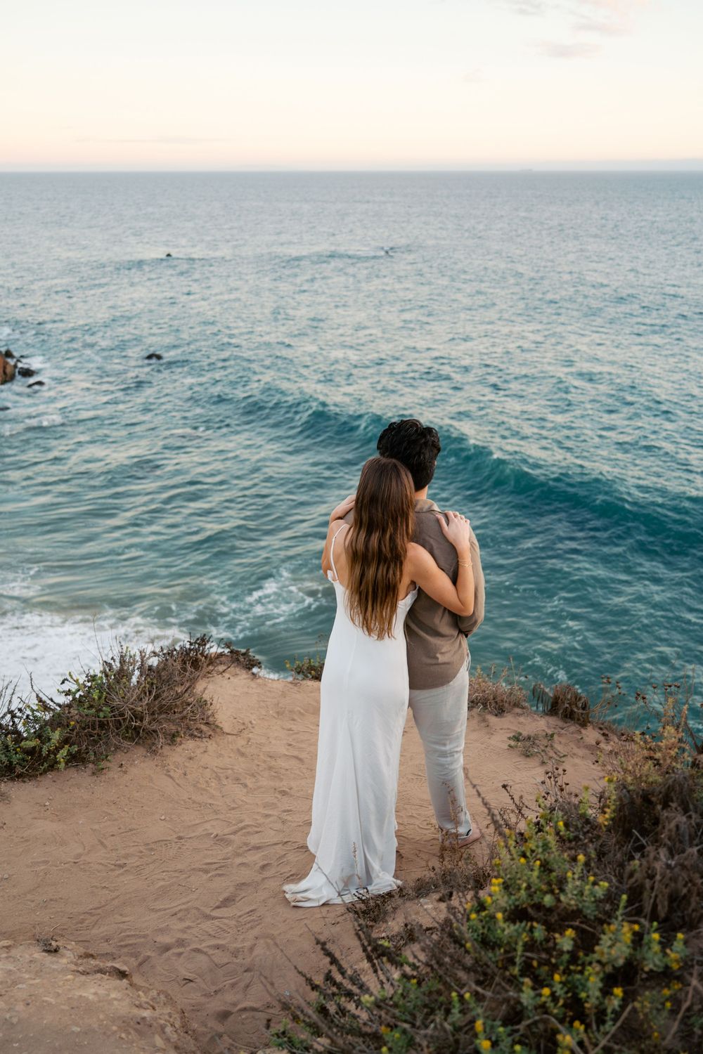Two people standing on a cliff edge overlooking the vast blue ocean during golden hour.