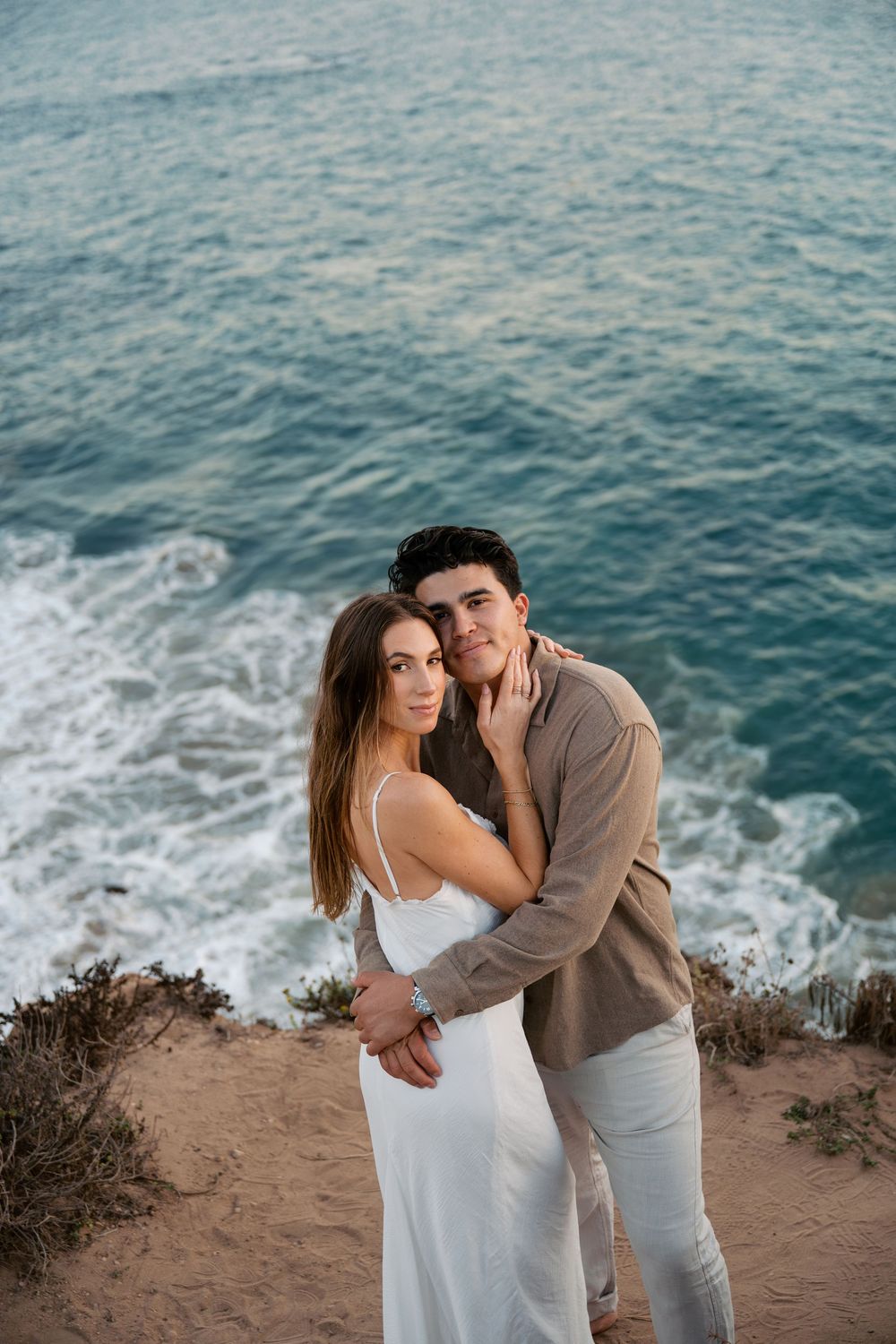 A romantic couple in white and beige outfits embrace on a coastal cliff overlooking the blue ocean waves.