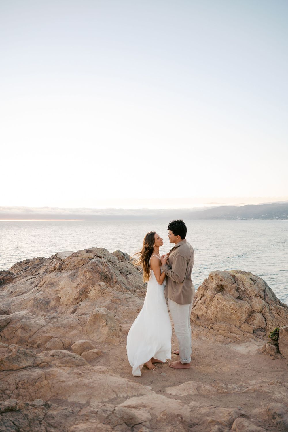 A couple in white and beige outfits embrace on rocky coastal cliffs during sunset.
