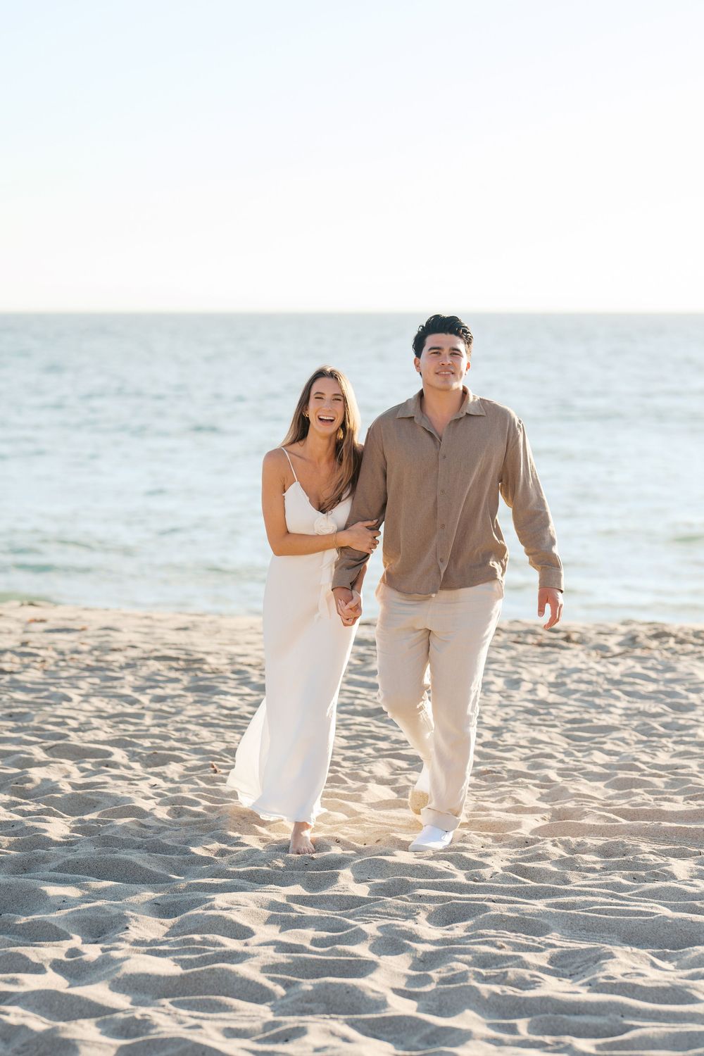 A couple walking together on a sandy beach wearing light colored casual clothing at sunset.