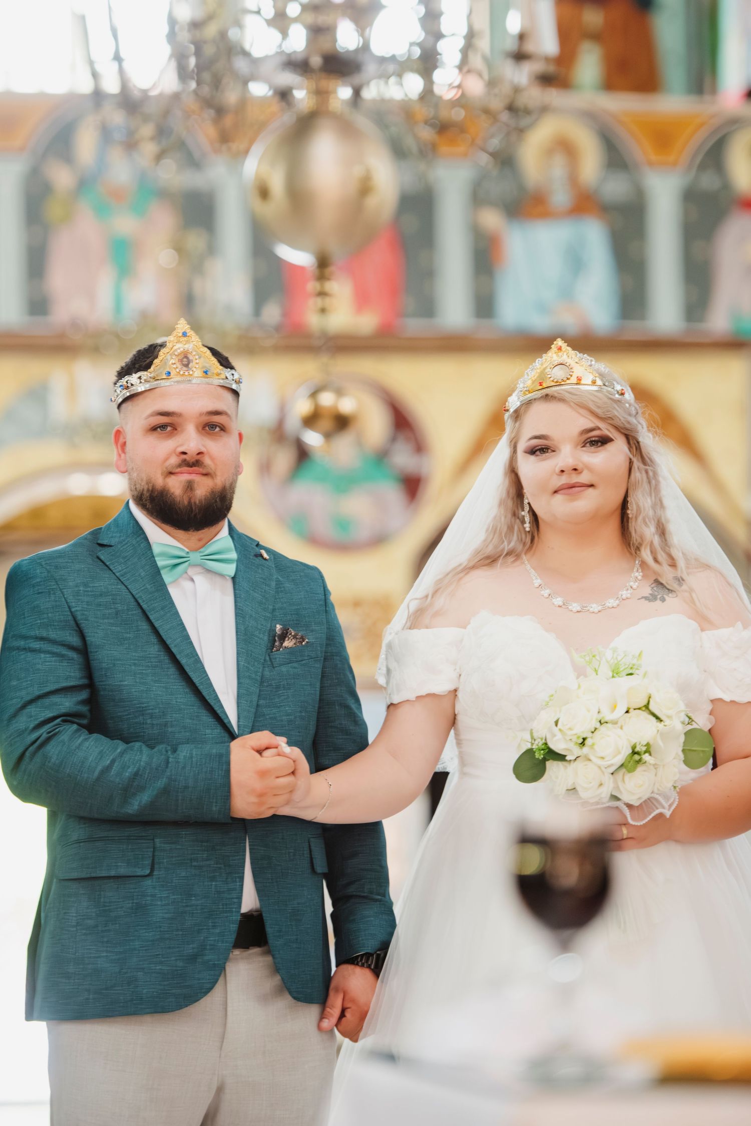 A bride and groom in formal wedding attire pose together in a carousel setting, with the groom wearing a teal suit jacket.