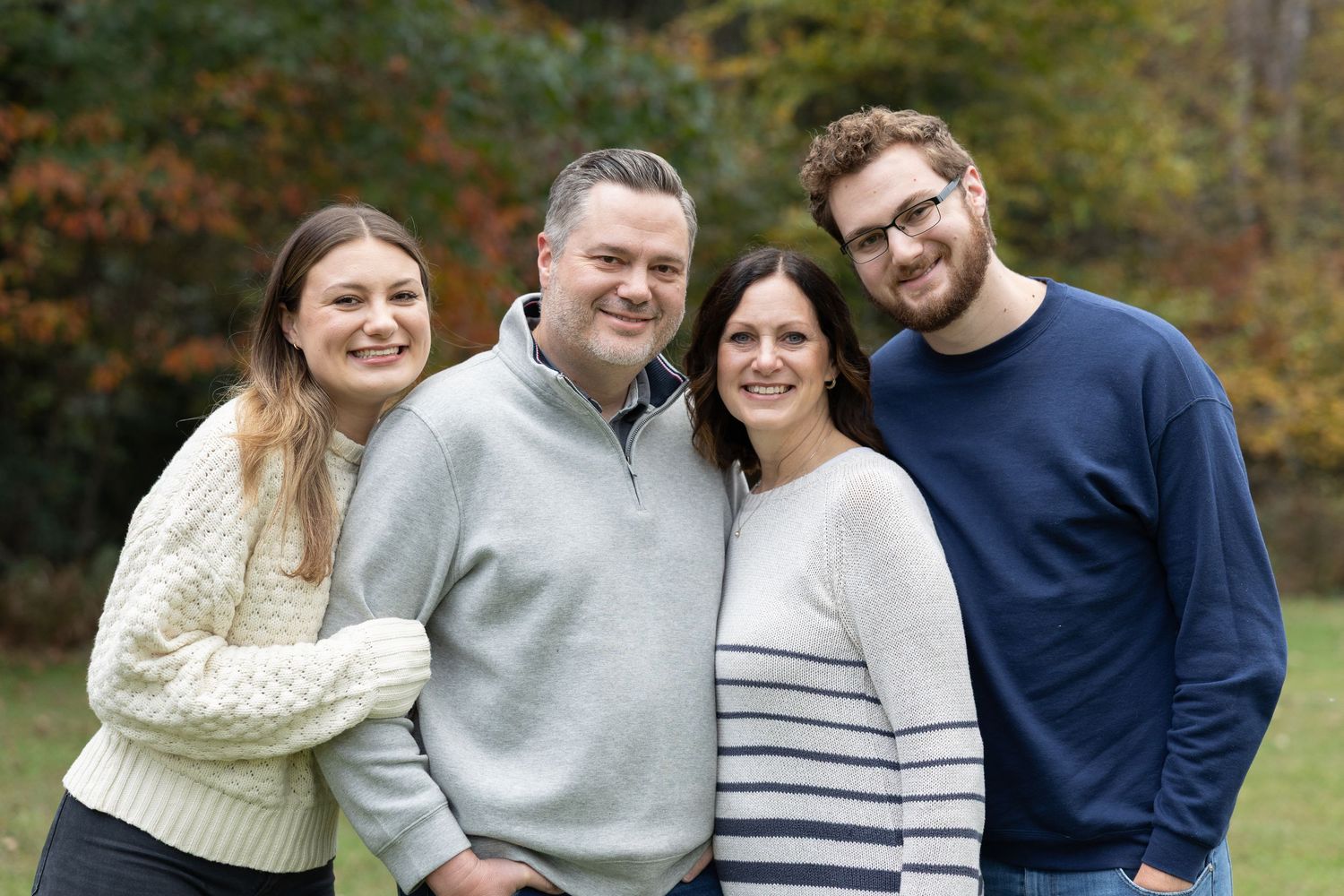 Smiling family of four posing together outdoors with colorful autumn foliage in the background.