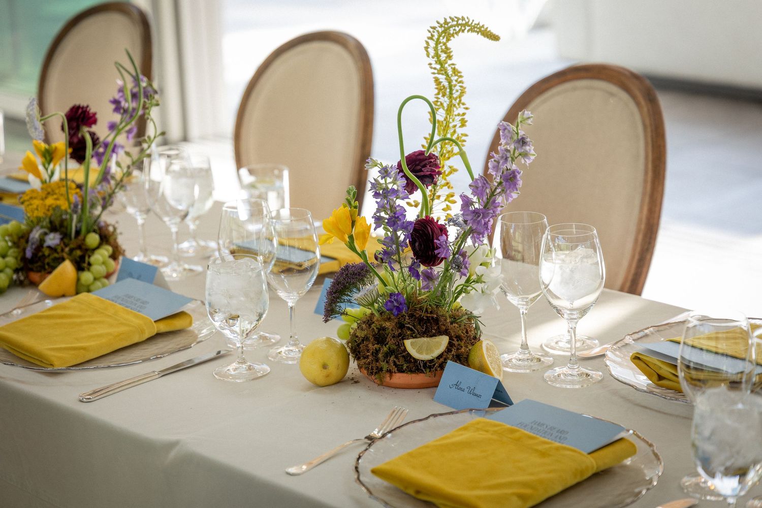 Elegant table settings with yellow napkins and purple wildflower centerpieces in rustic dining room.