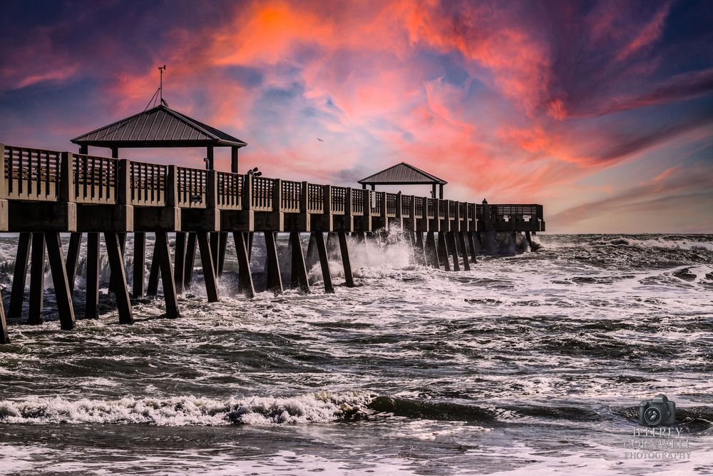 Dramatic sunset illuminates a wooden fishing pier extending into choppy ocean waters.