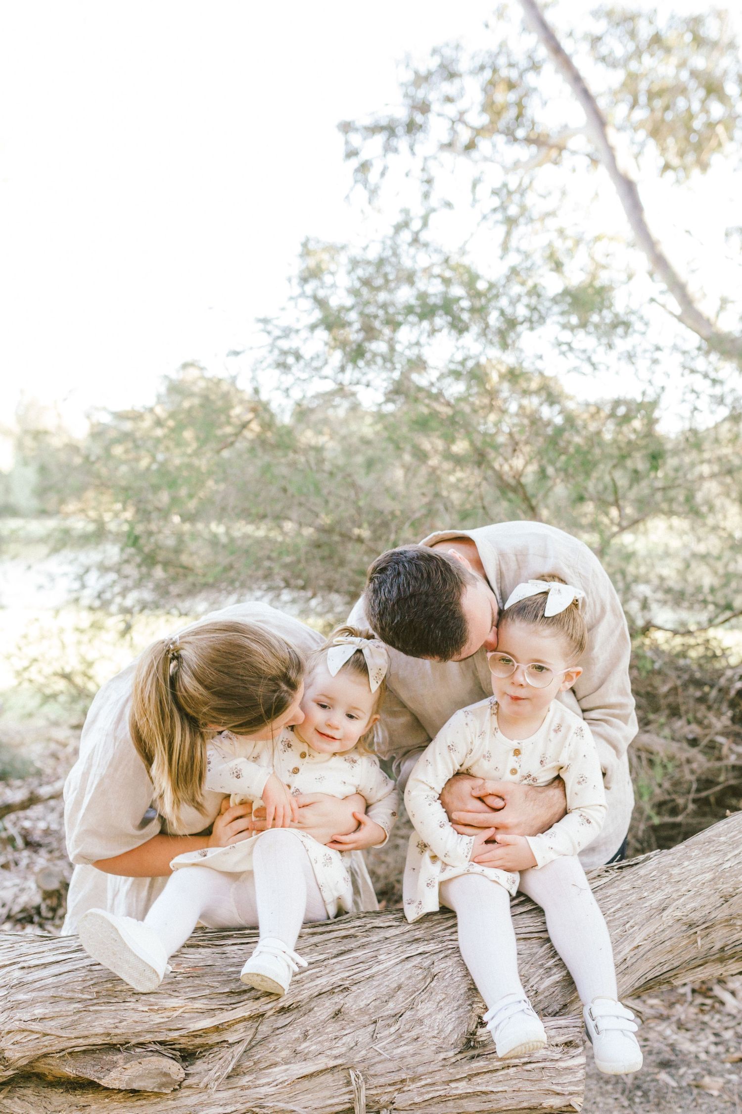 A sequence of four sweet family moments captured on a fallen tree in natural lighting wearing matching white outfits.