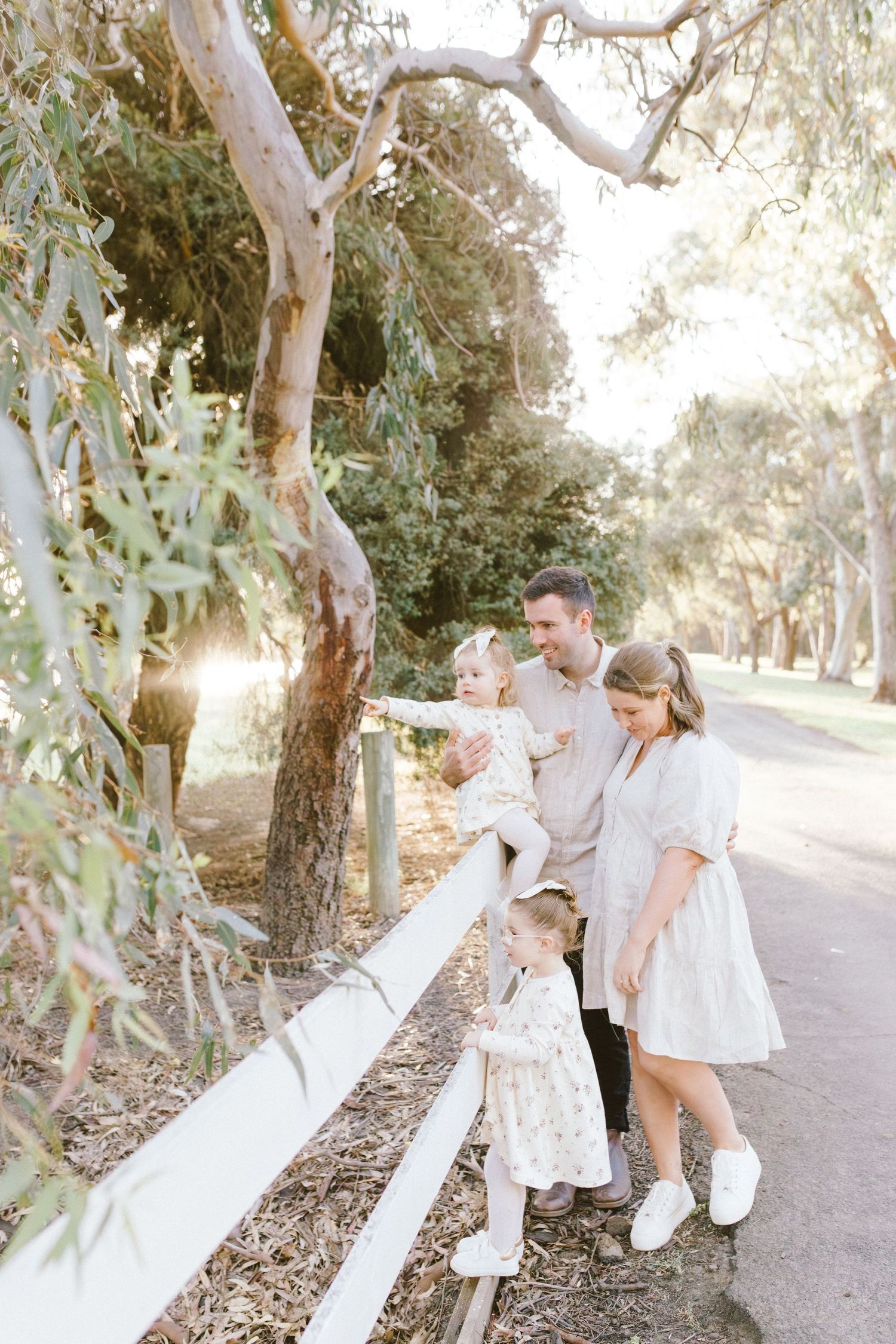 A loving family dressed in white enjoys playful moments along a tree-lined path during golden hour.