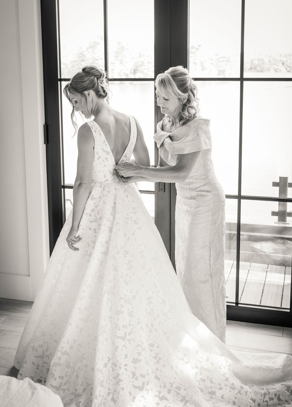 A tender black and white moment of wedding dress preparations by a large window.
