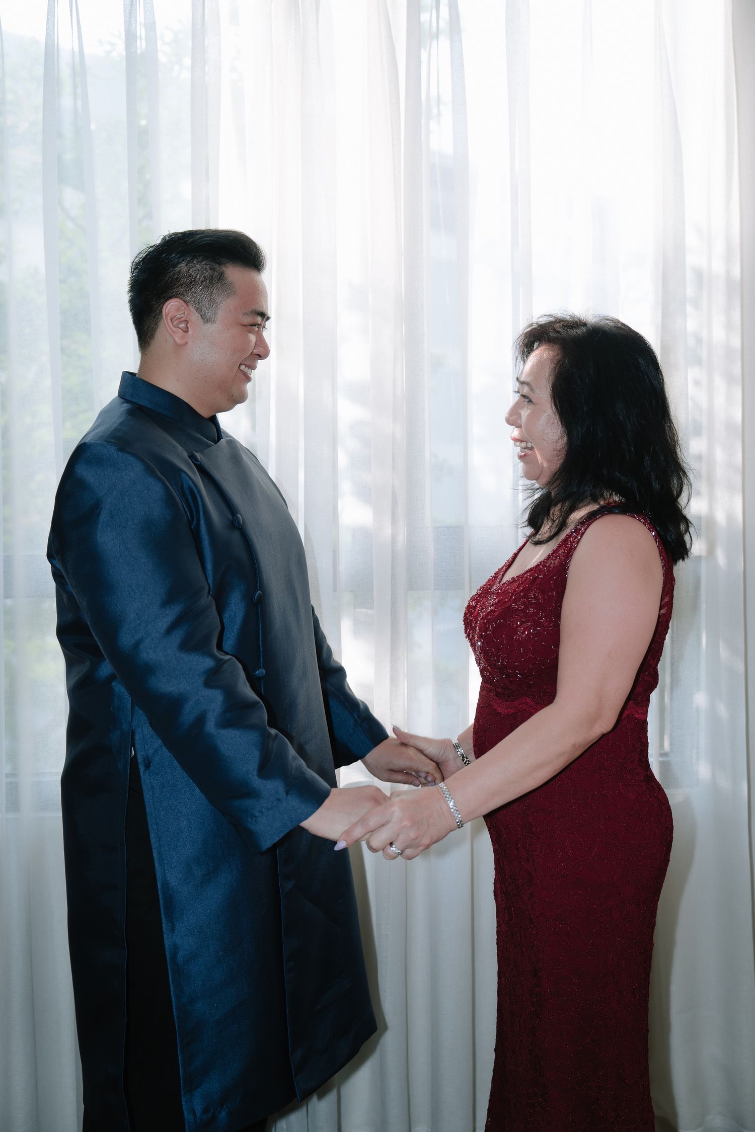 Groom in blue mandarin suit holding hands with mother in red dress by window.