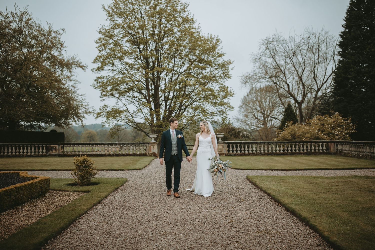 Couple strolls hand in hand along a tree-lined gravel path on an autumn day at a historic estate.