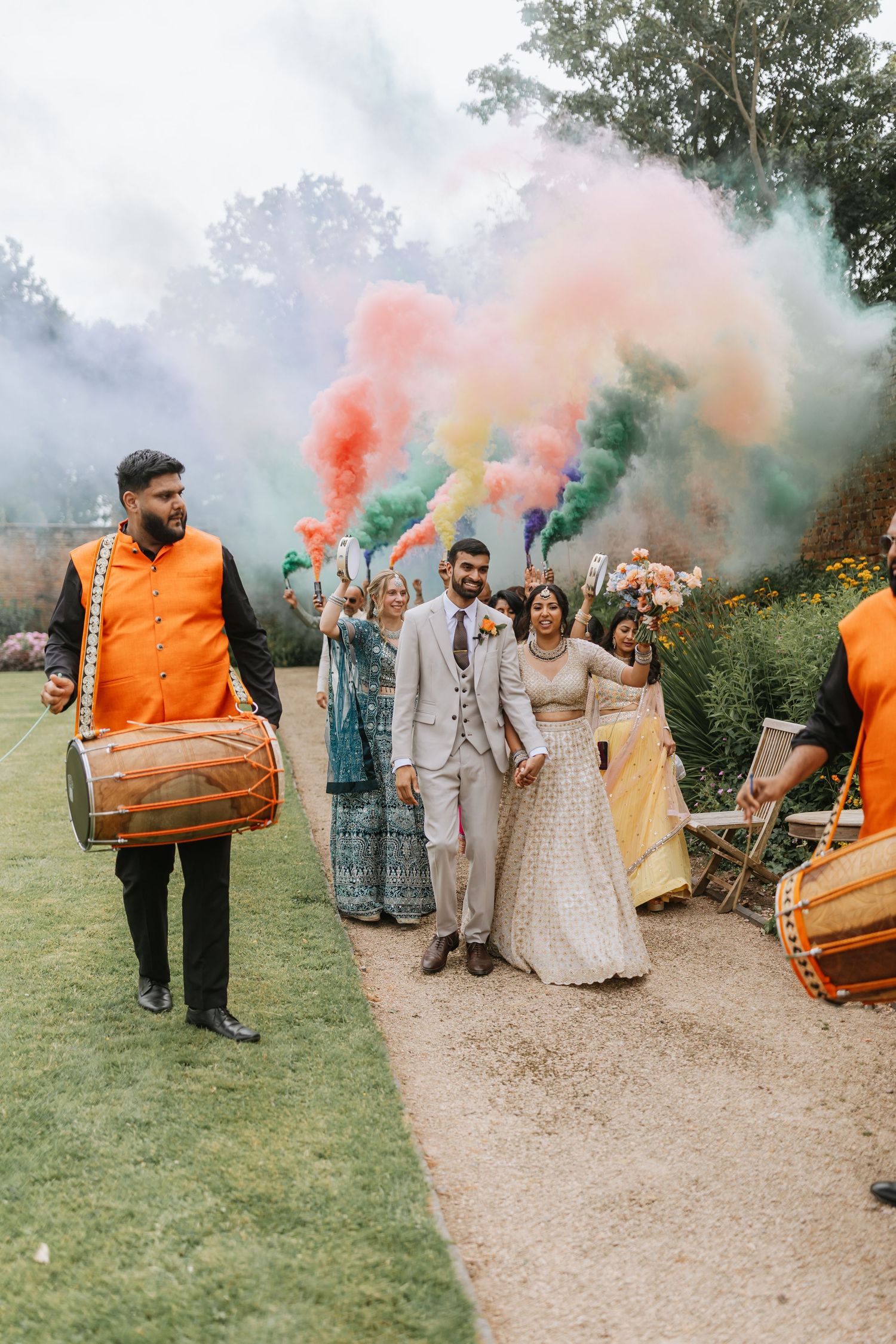Colourful smoke bombs create a vibrant outdoor celebration as wedding guests gather on a garden path. Thorpe Garden Wedding.