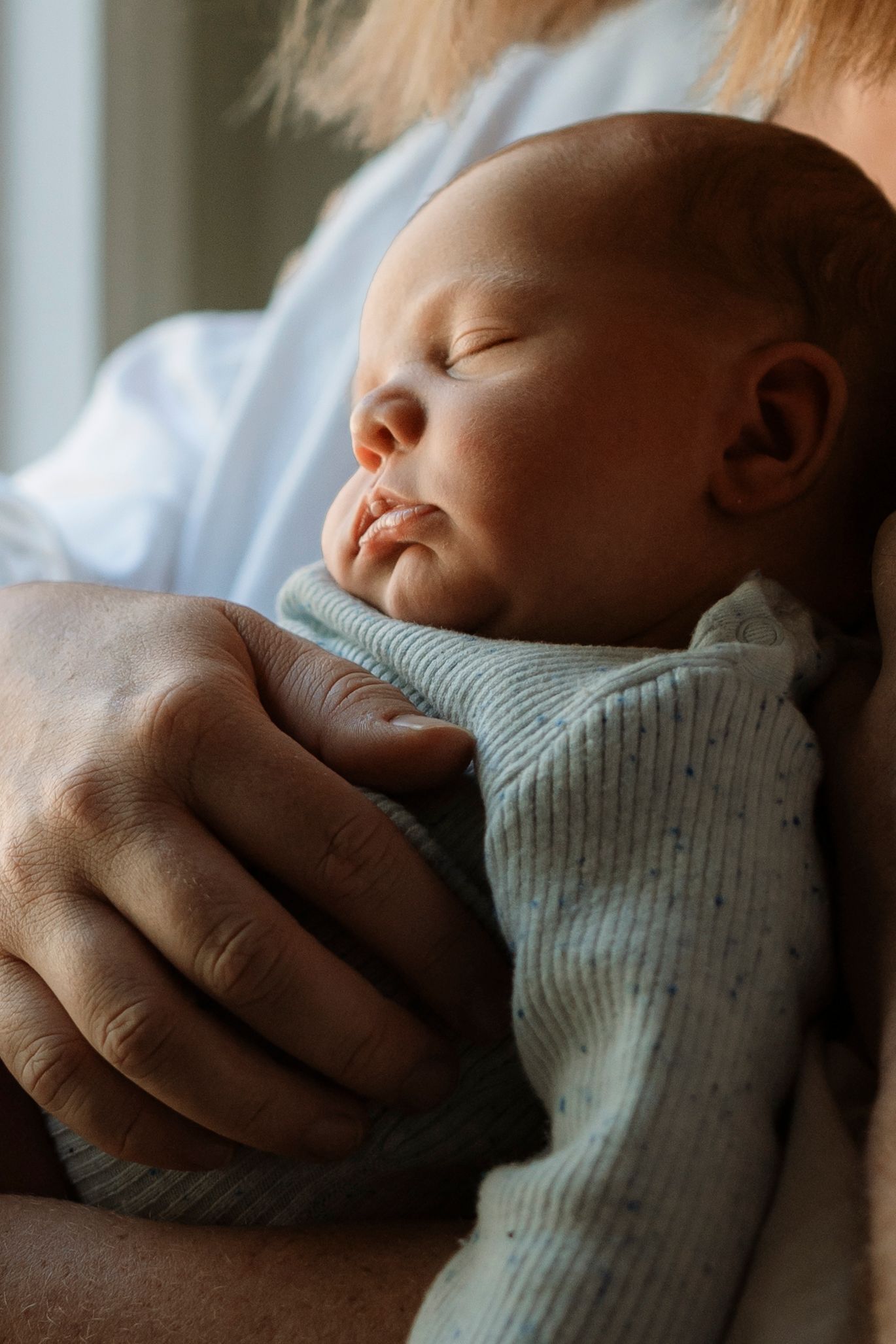 A peaceful sleeping baby wearing a light blue ribbed sweater is cradled in gentle hands.
