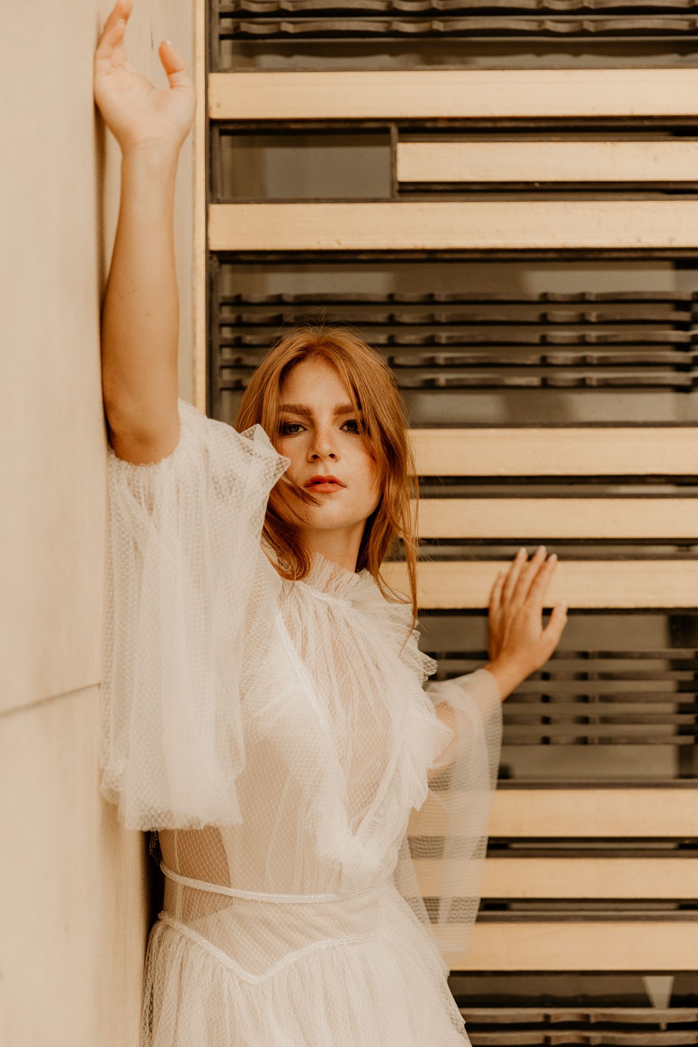 Model in an ethereal white dress poses in front of window blinds.