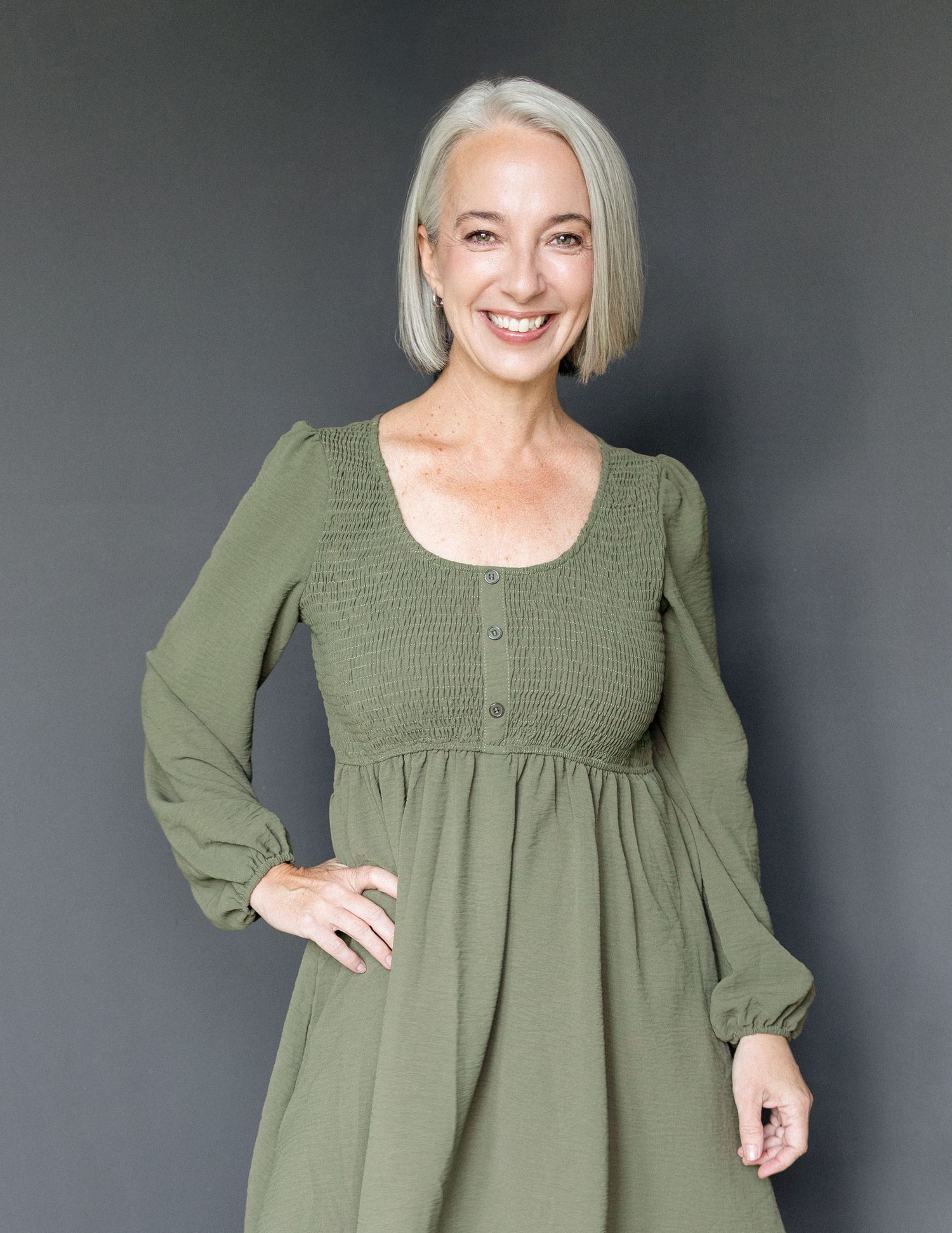 Smiling woman with short silver hair wearing green dress with buttons and hand on hip on a dark grey backdrop.