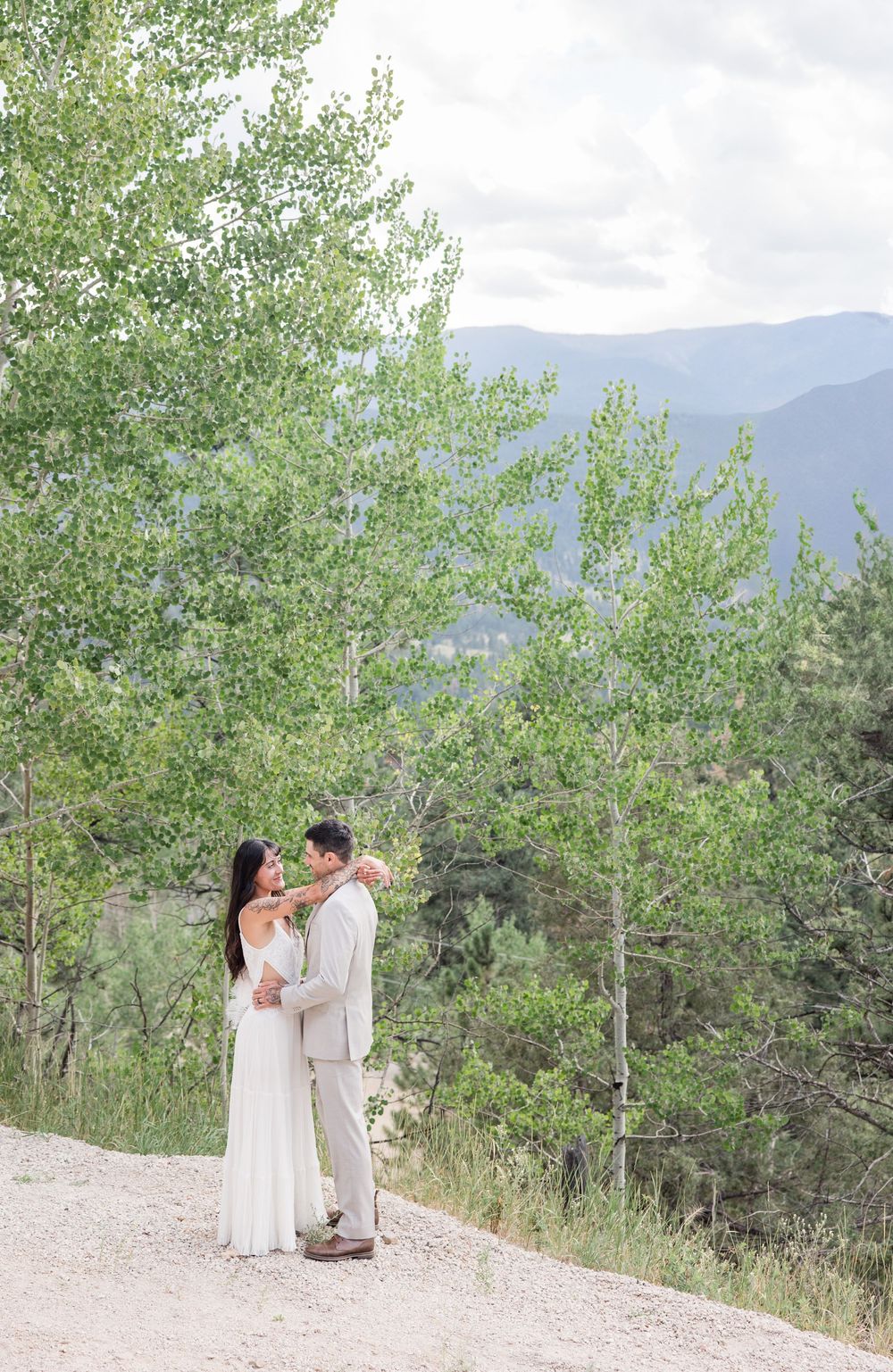 Wedding couple in white dress and beige suit embracing on a scenic mountain overlook with trees.