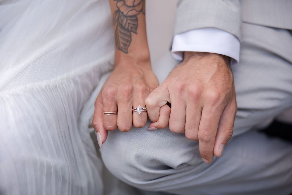 Close-up view of bride and groom's hands with intertwined fingers showing wedding rings and tattoos.