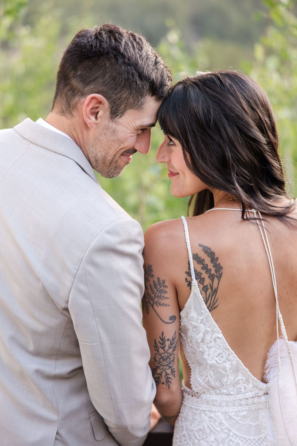 Intimate moment between couple showing bride's intricate back tattoos in outdoor wedding setting.