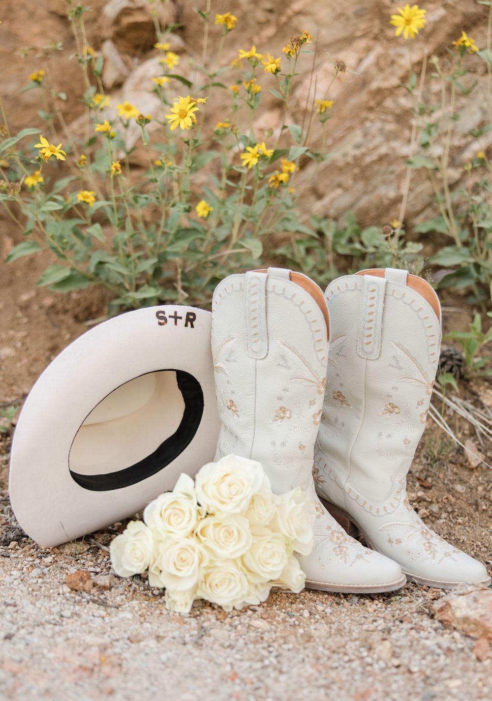 White cowboy boots and hat with white roses displayed against yellow wildflowers in a rustic outdoor setting.