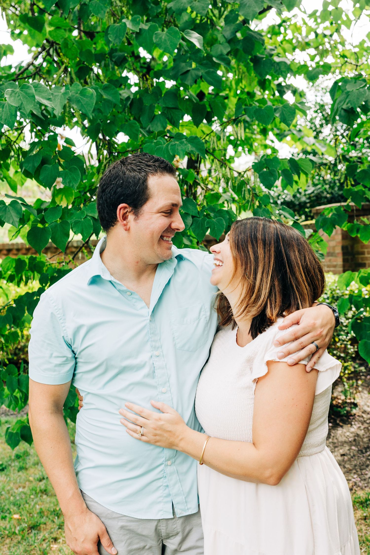 A romantic moment captured under green leafy trees as a couple embraces in casual summer attire.