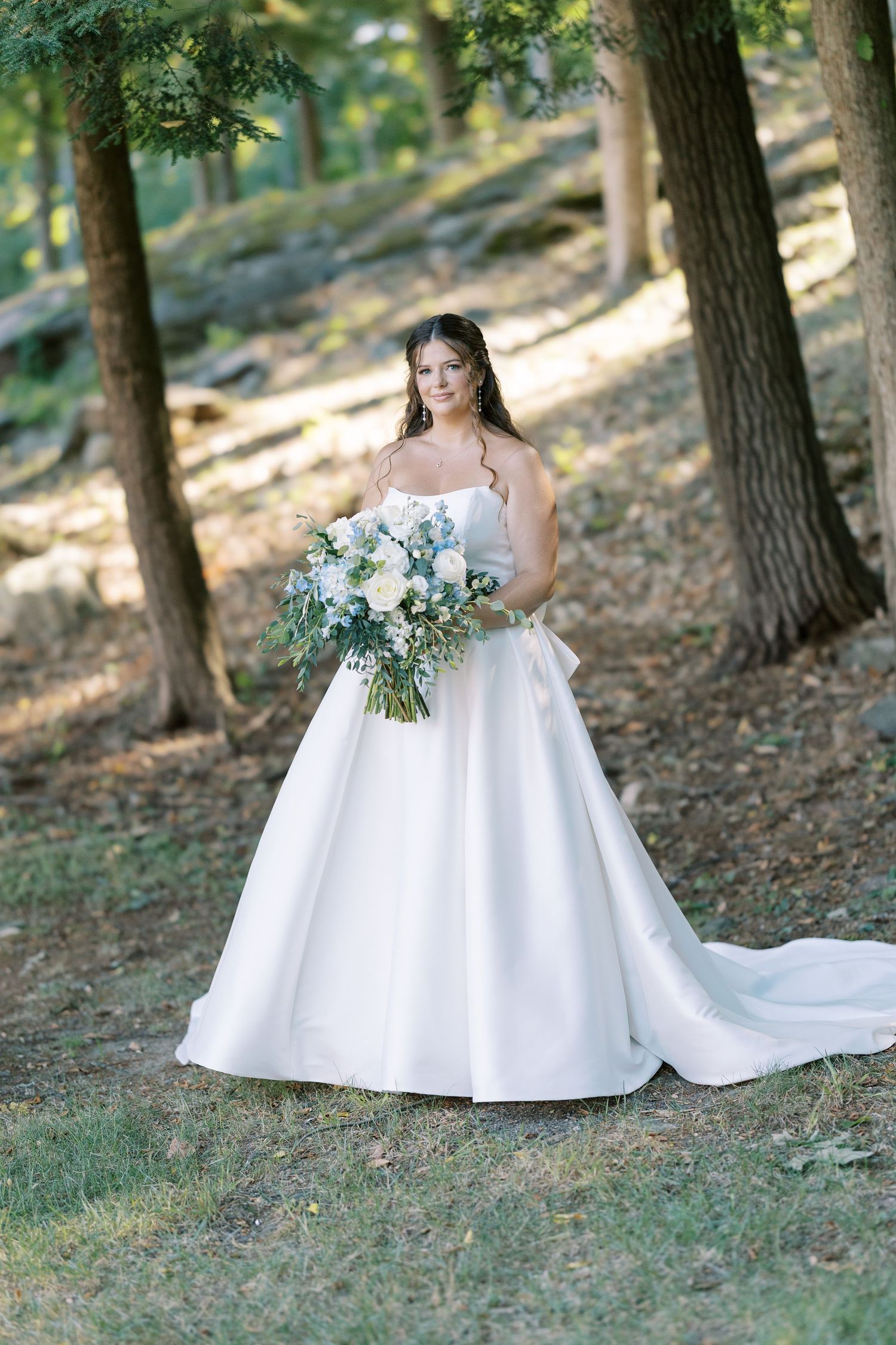 A bride in a white ballgown poses with a bouquet of blue and white flowers in a sunlit wooded forest setting.
