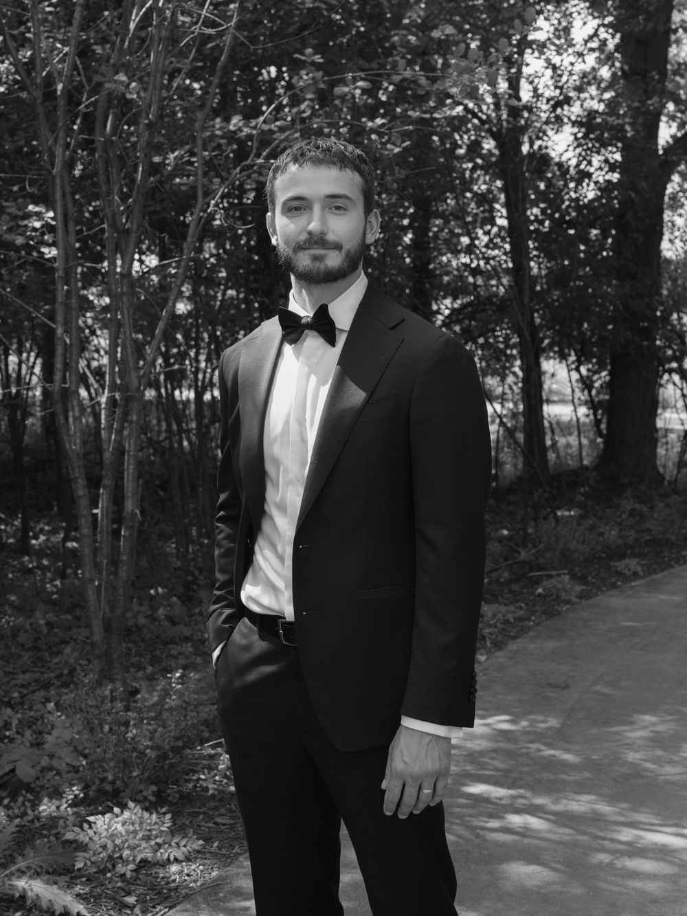 Formal portraits of someone wearing a black tuxedo and bowtie in an outdoor wooded setting.
