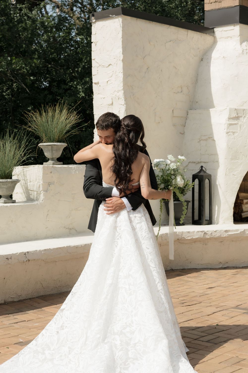 Two people in elegant white wedding dresses share intimate moments on a stone patio with white walls and greenery.