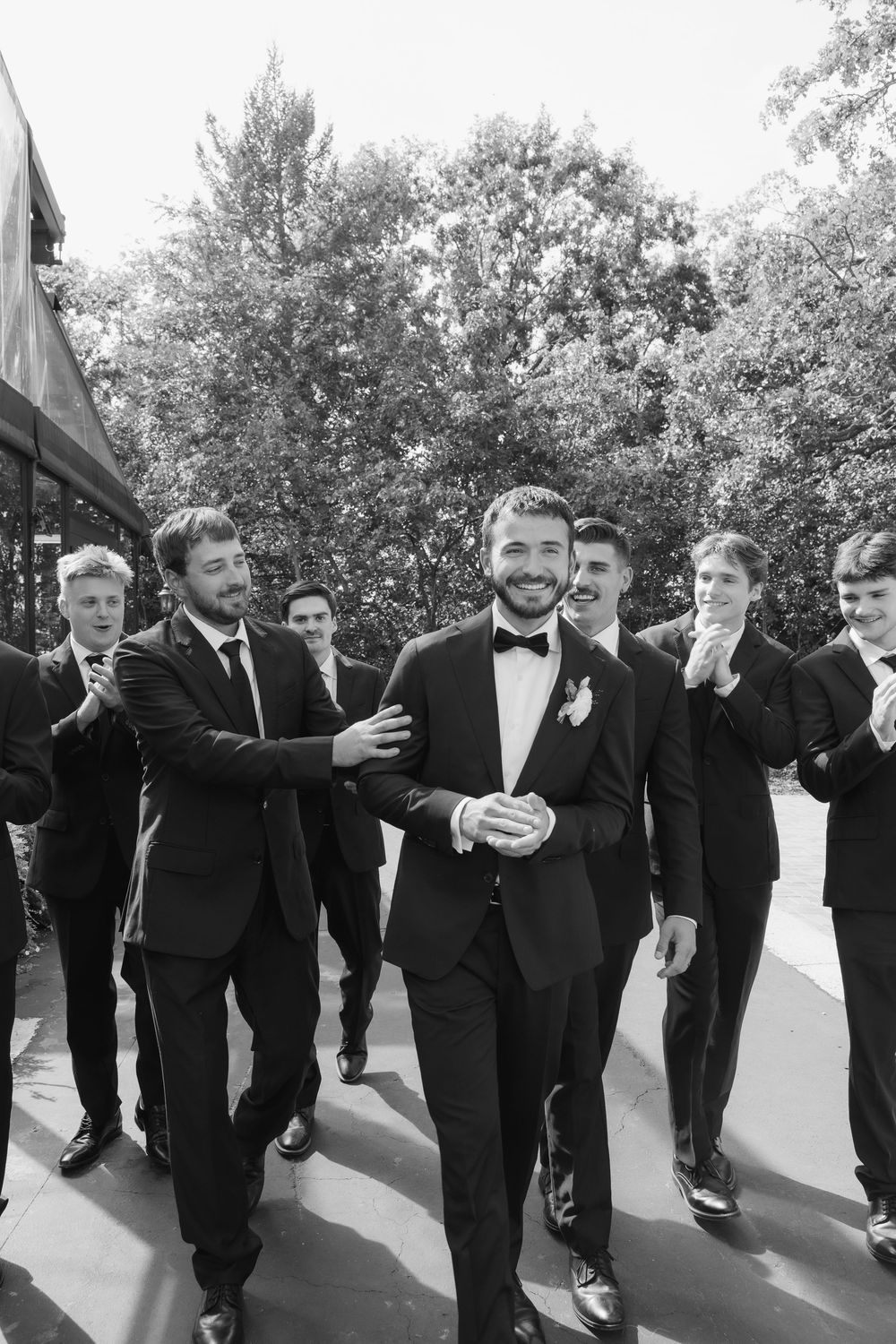 A line of groomsmen in black tuxedos pose together outdoors while walking in sequence.