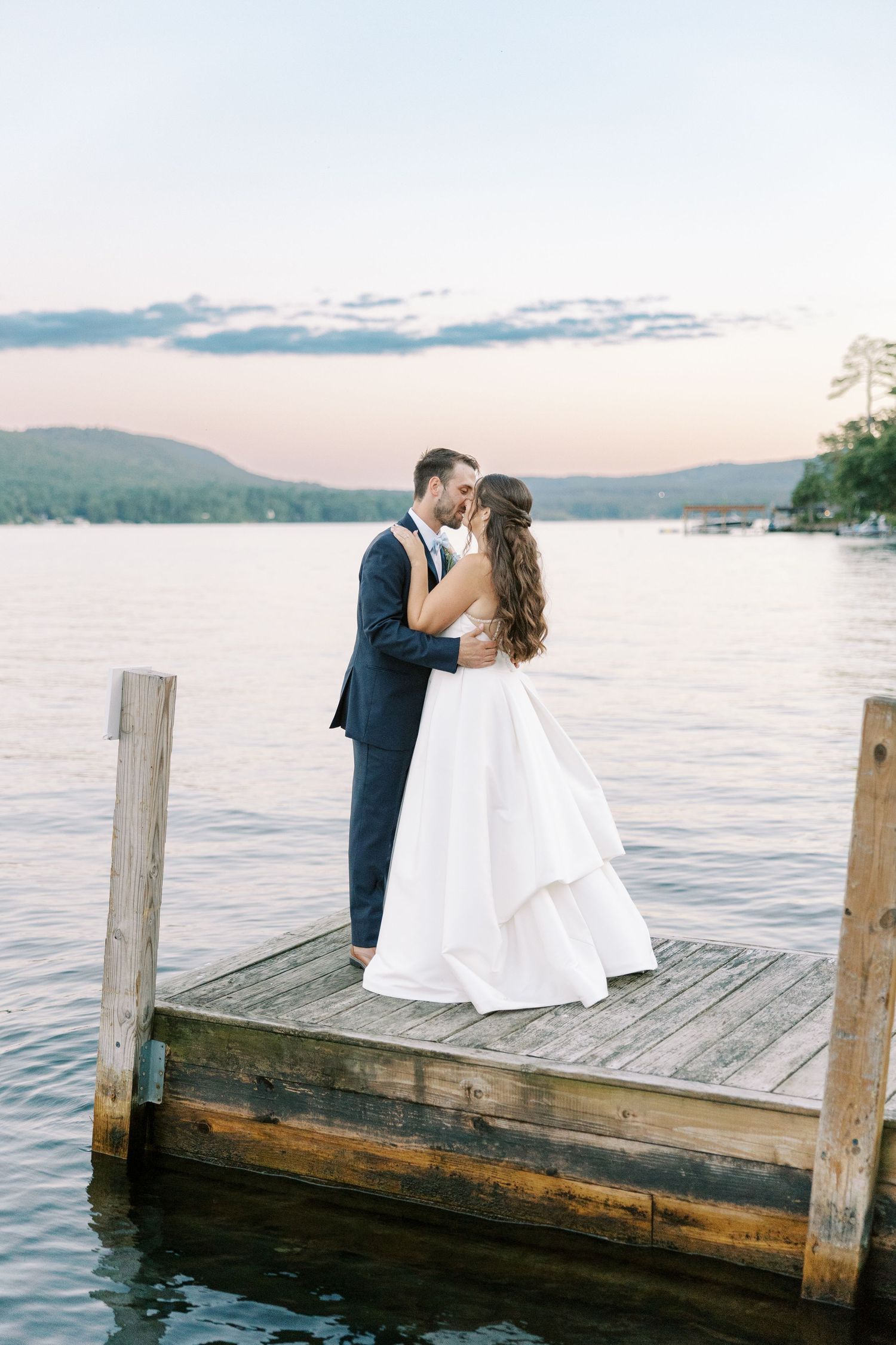 A couple shares a romantic kiss on a wooden dock overlooking a serene lake at sunset.