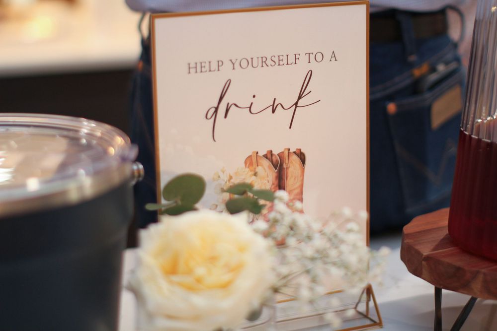 An elegant wedding reception sign reading 'Help yourself to a drink' displayed next to a white rose.