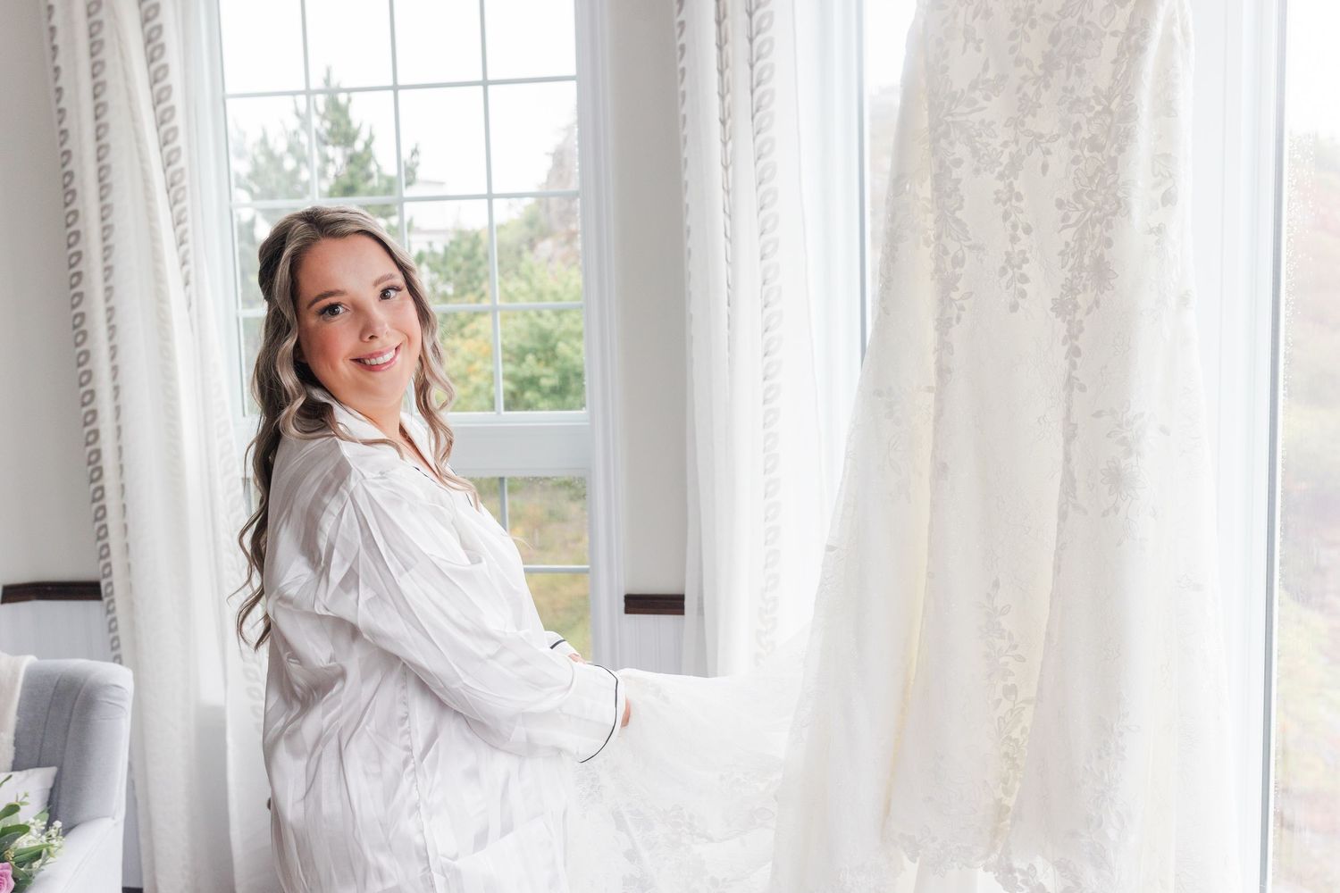 A light-filled bedroom with white curtains and windows features someone standing in a flowy white robe.