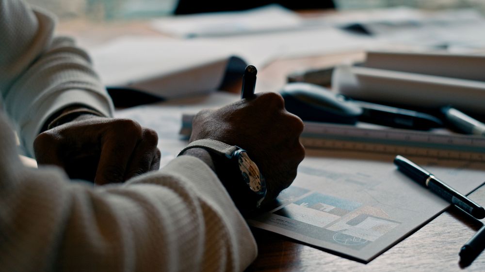 Close-up view of hands writing on paper with pens and office supplies on desk.
