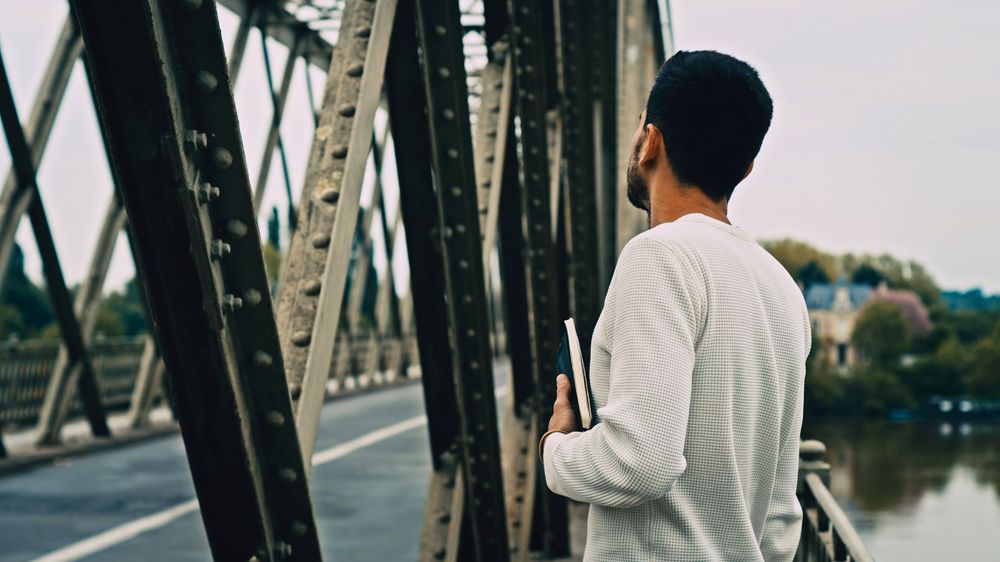 Person in white shirt looking at industrial metal bridge over water.