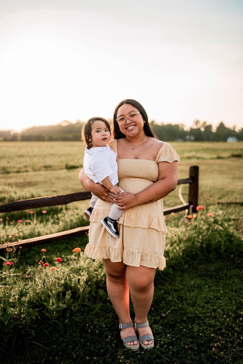 A serene family moment captured at sunset by a wooden fence surrounded by wildflowers in a rural field.