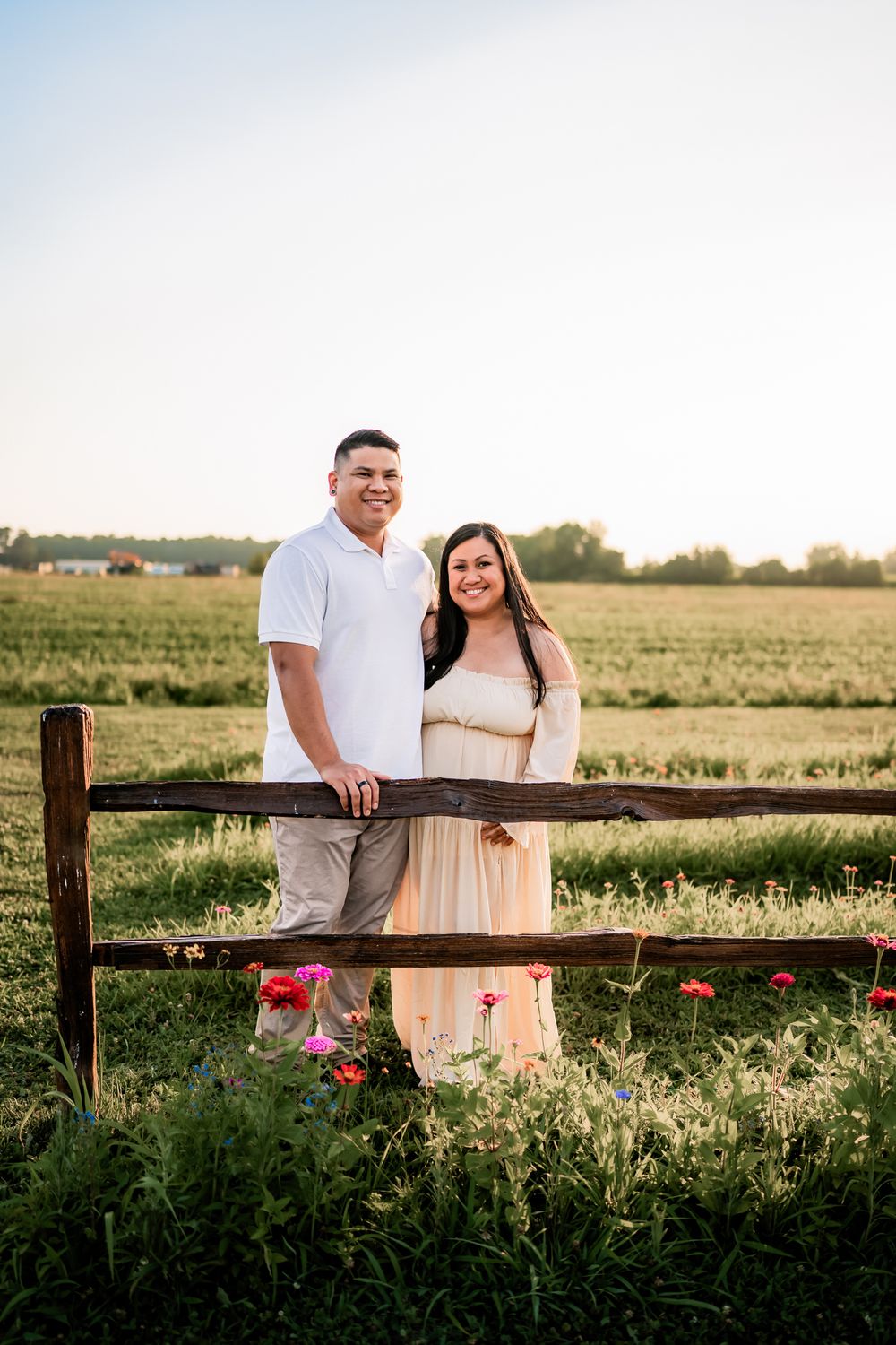 Couple stands together at rustic wooden fence in golden sunlit field during sunset photoshoot.
