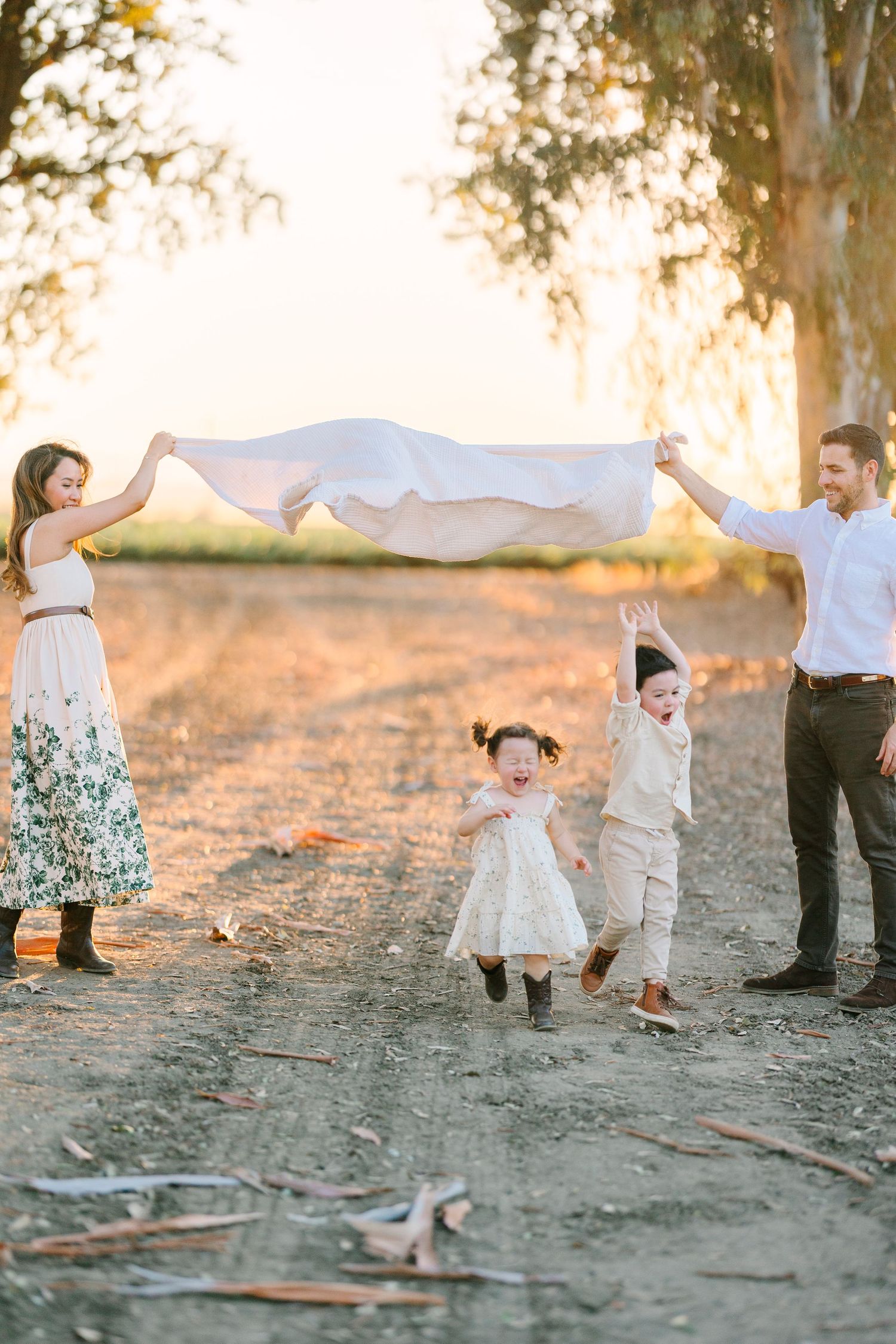 A family plays together under a white sheet during golden hour in a rustic outdoor setting.