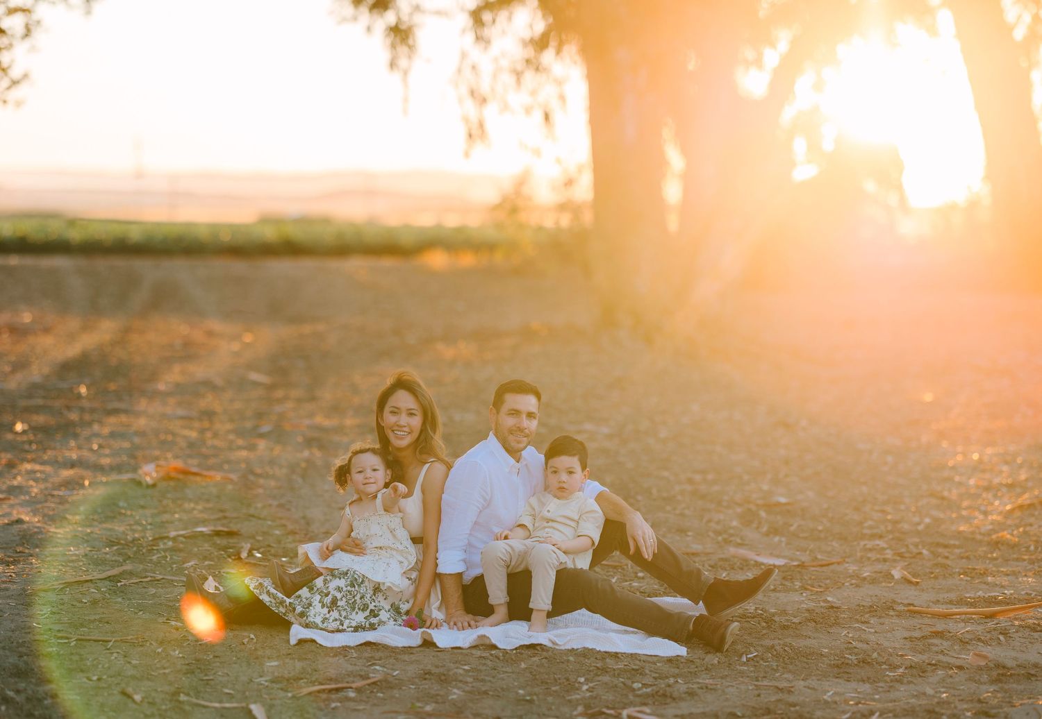 A family relaxes together on a blanket during sunset with trees in the background.