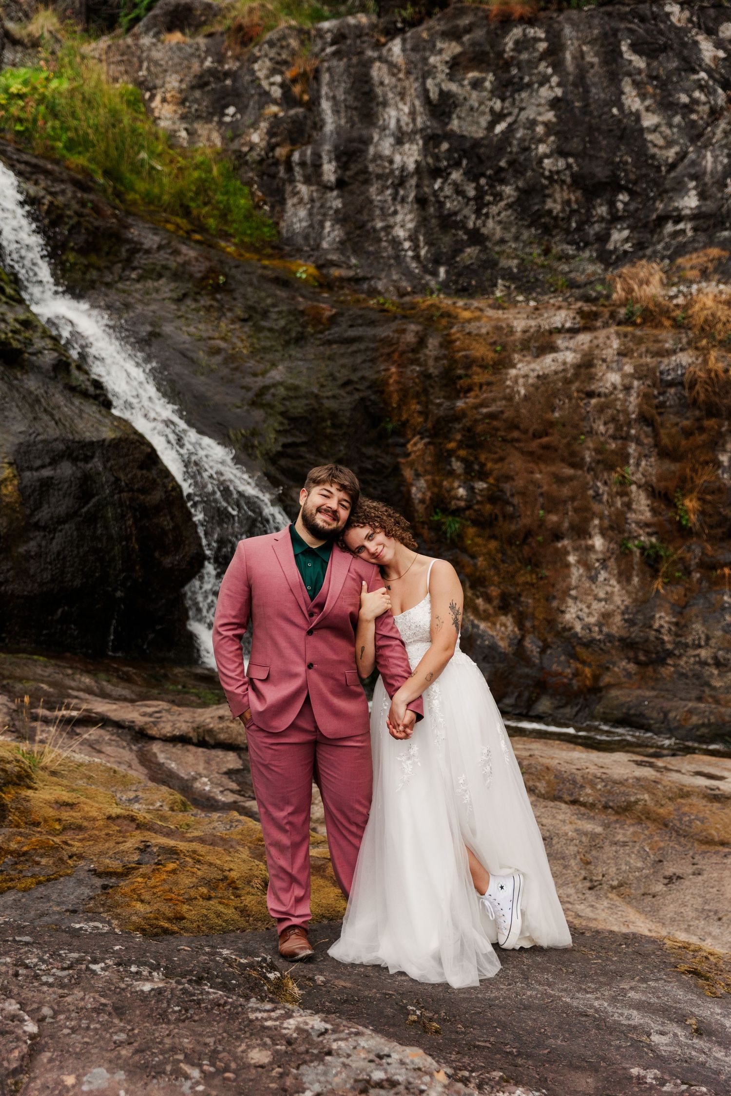 Couple poses together against rocky cliff with cascading waterfall in background for outdoor wedding photos.