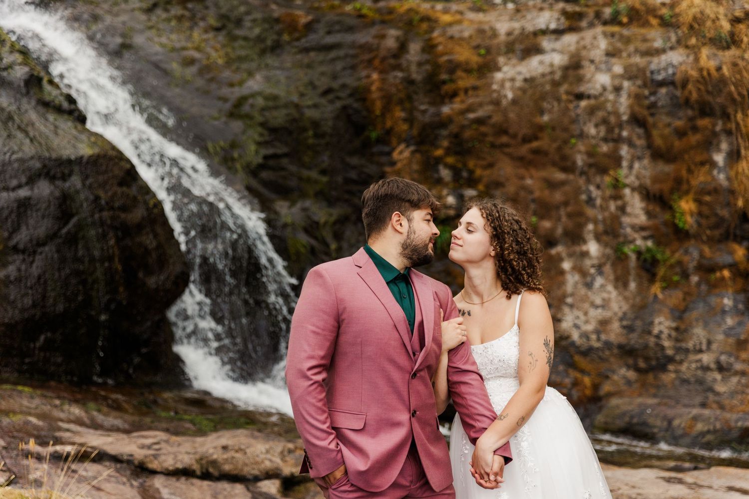 A romantic couple dressed in formal wear stands close together in front of a cascading waterfall against rocky cliffs.