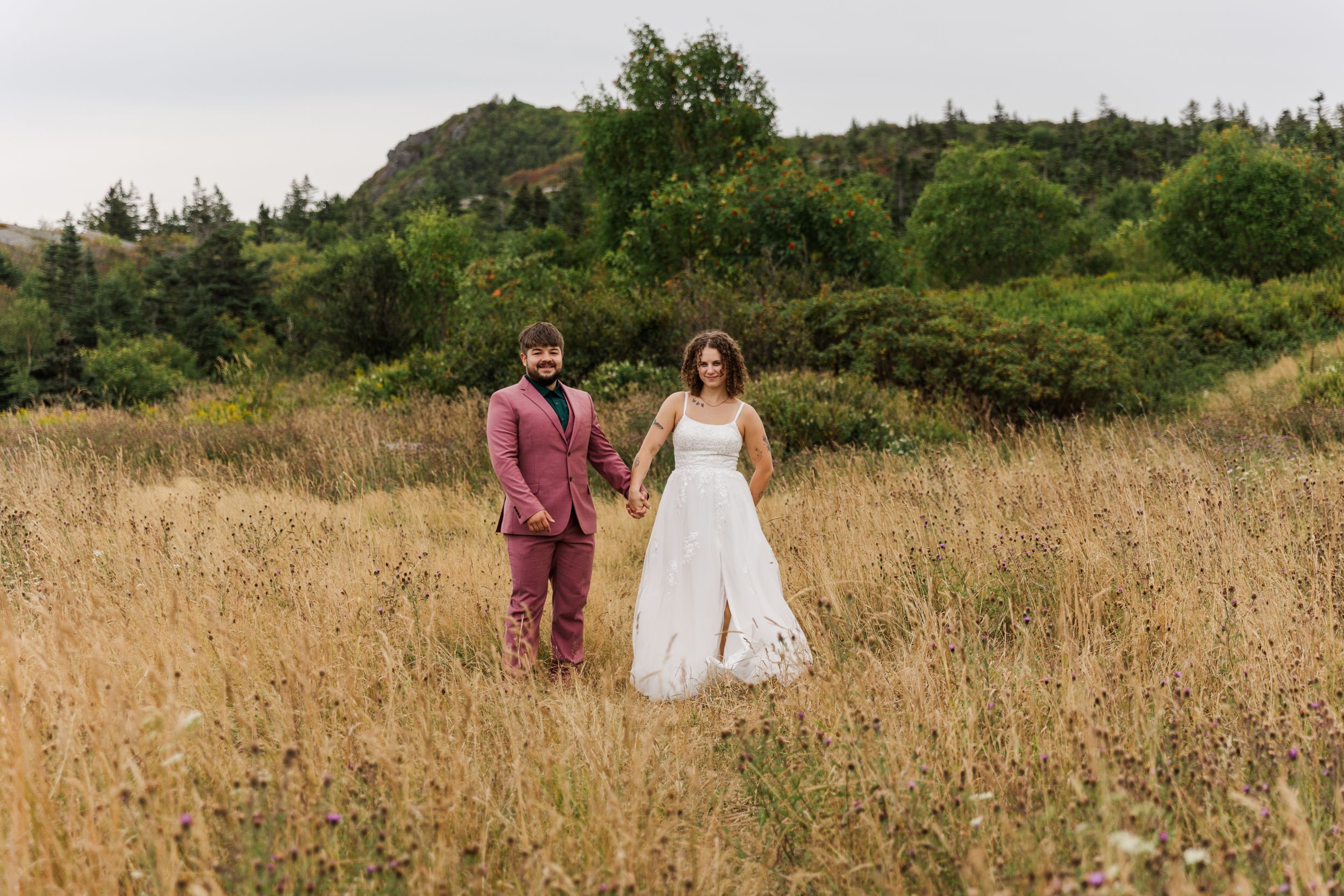 Two people walking through tall golden grass with mountains and trees in the background on a summer day.