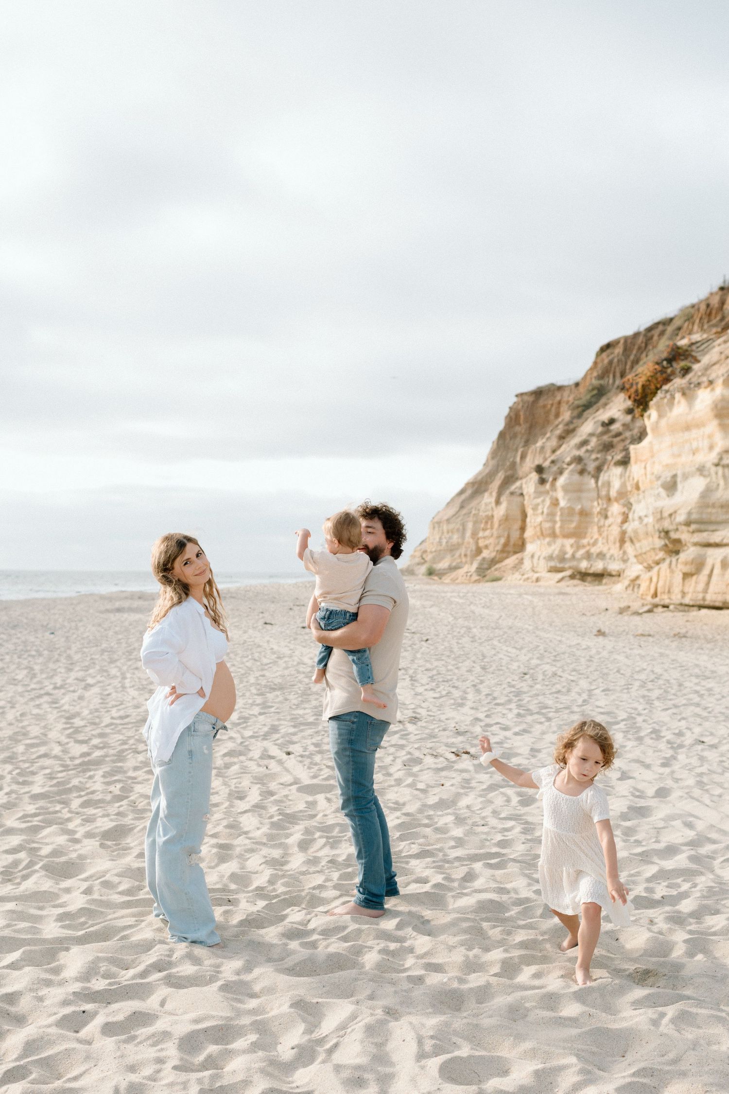 A family in light colored outfits dances and plays together on a sandy beach with towering cliffs in the background.