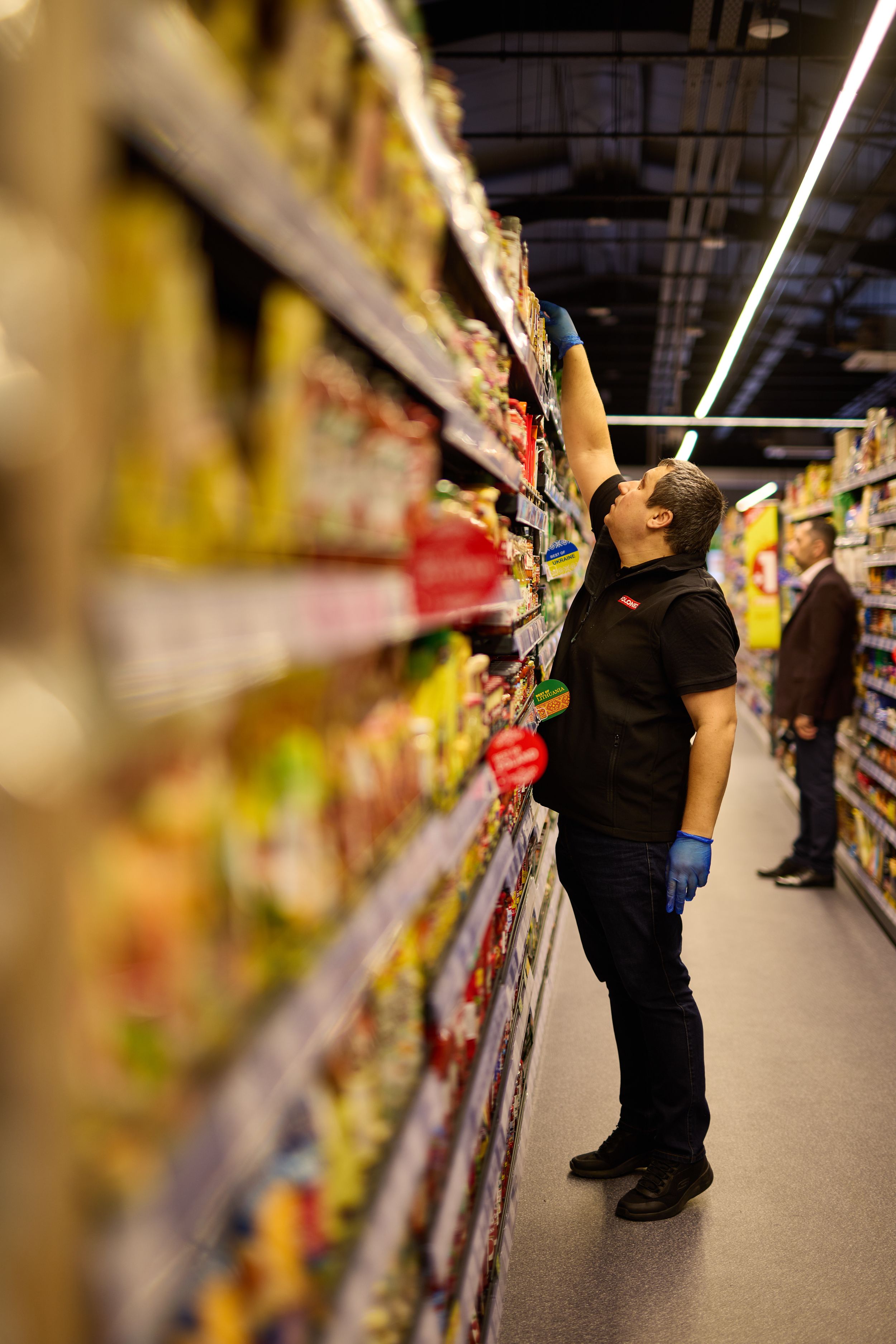 Employee reaching for products on high supermarket shelf.