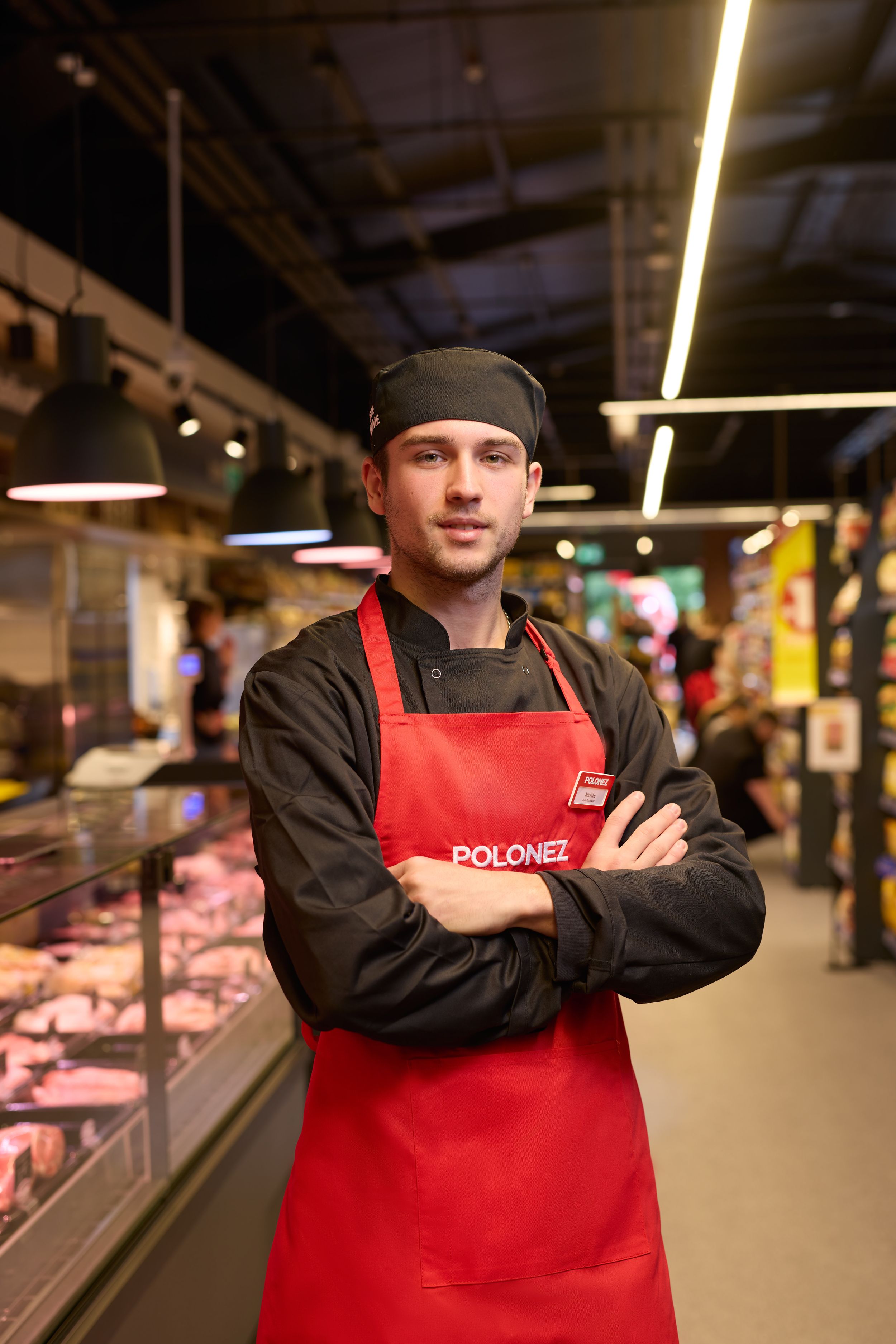 Supermarket worker in red apron standing by meat counter.