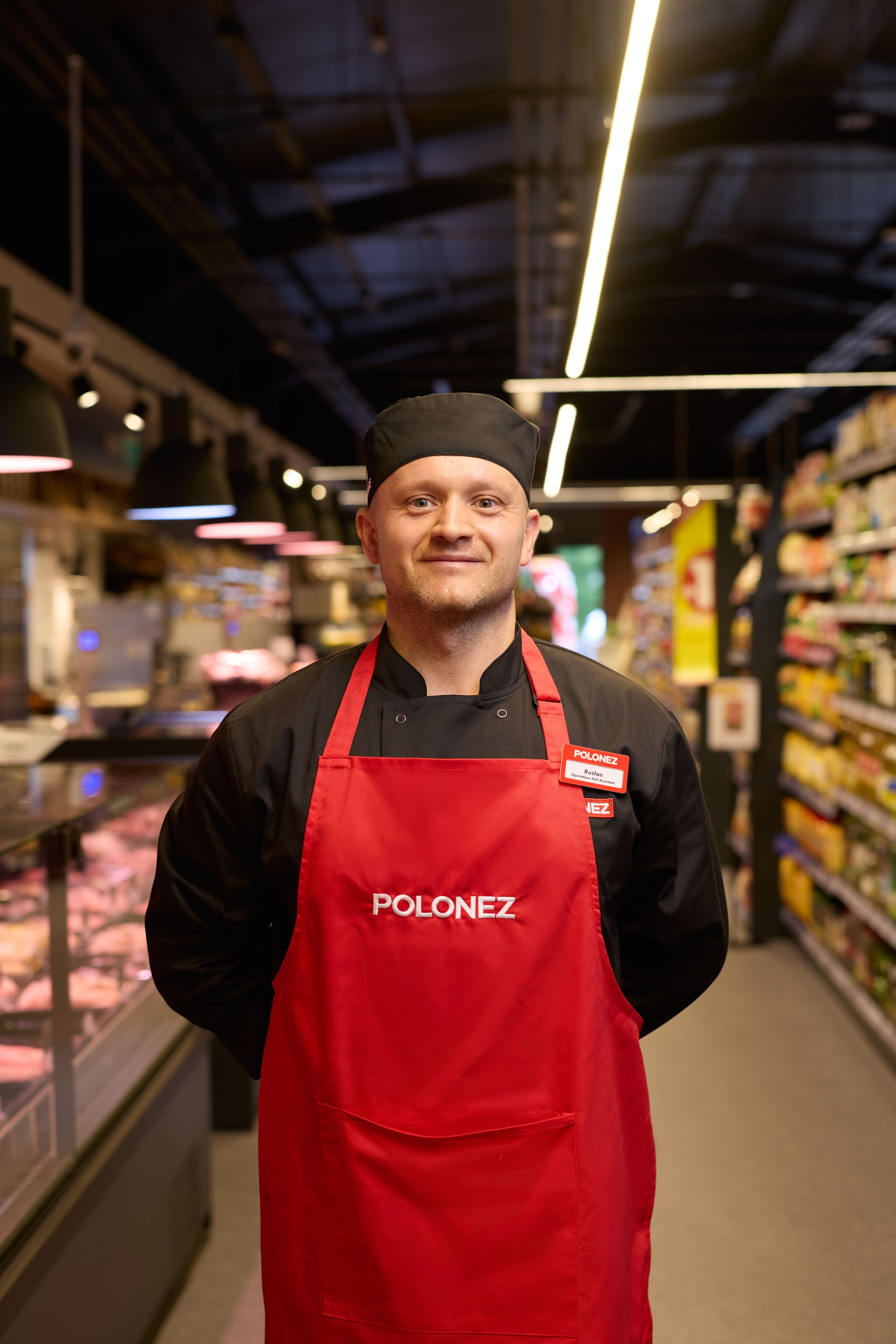 Store employee wearing red Polonez apron and black uniform standing in supermarket aisle.