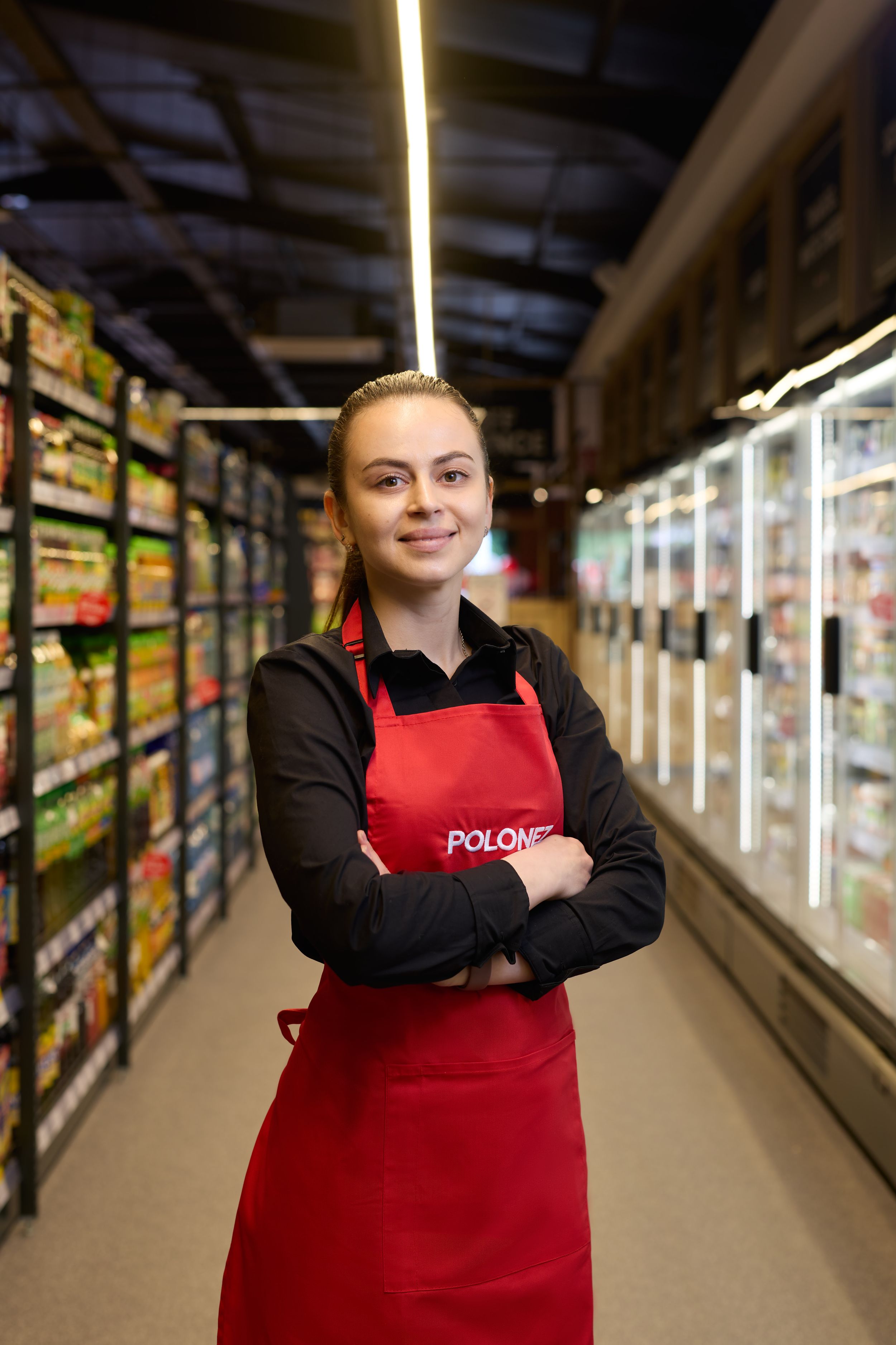 Staff member in red apron poses confidently in grocery store frozen food section.