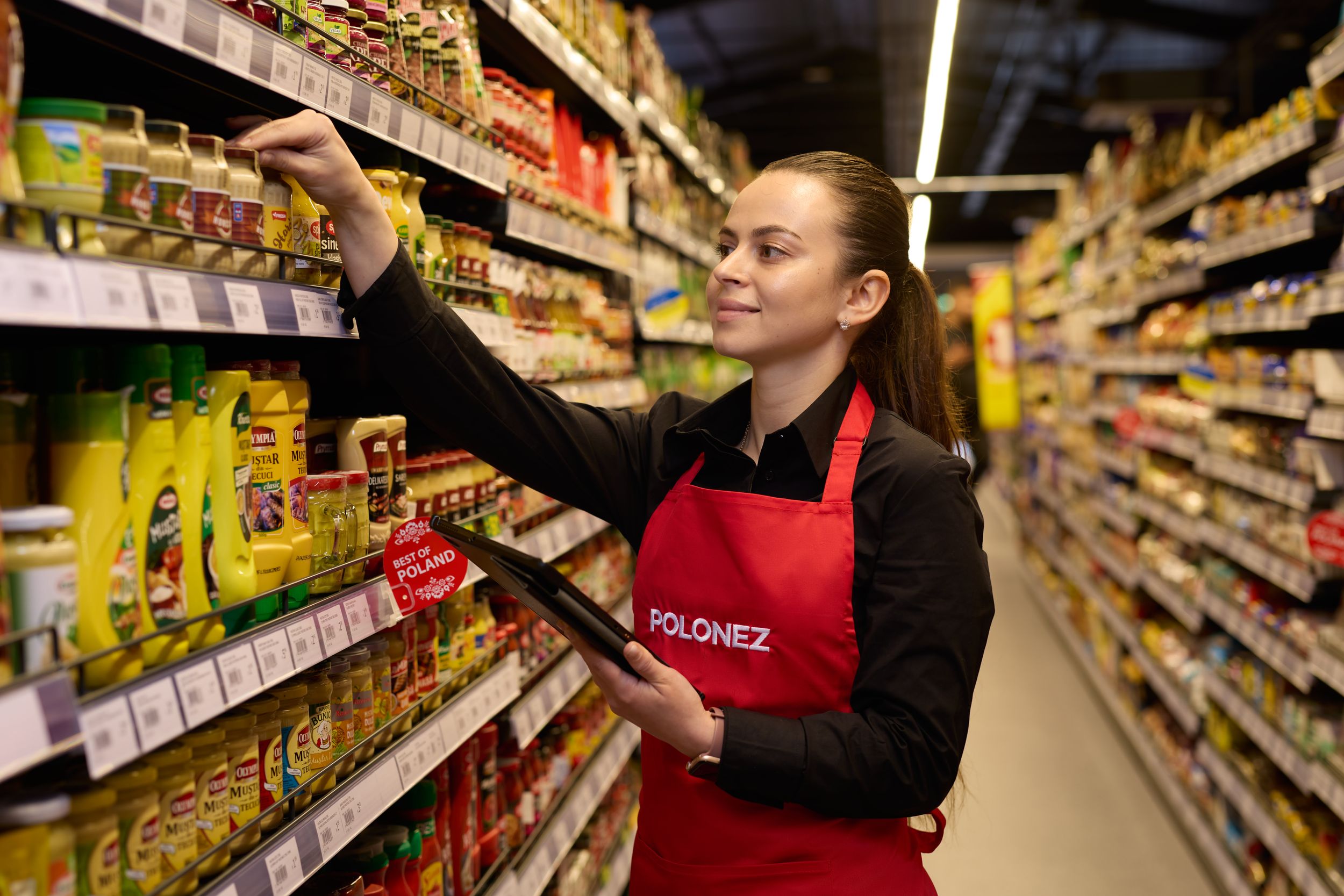 Employee stocking shelves in supermarket aisle wearing red apron.